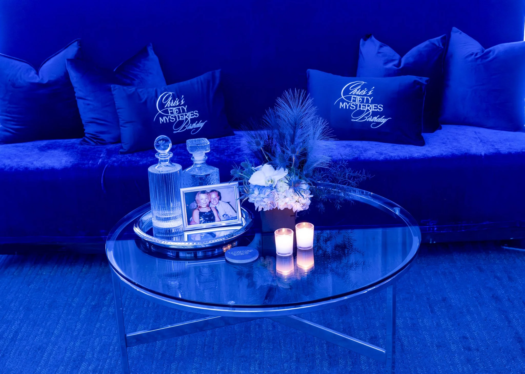 Living room with a glass coffee table, blue velvet sofa, and pillows that say "Christ's Fifty Mysteries Birthday." The coffee table holds a silver tray with a framed photo of a smiling couple, two glass decanters, a floral arrangement, and two lit candles. The room is illuminated with blue lighting.
