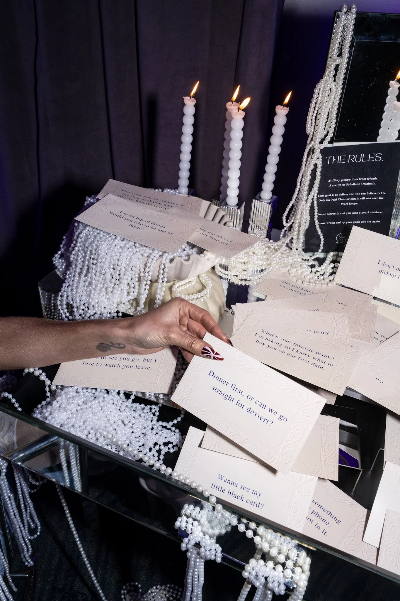 A person holding a white card with a red and white pattern, surrounded by multiple cards with questions and messages, on a table decorated with pearl necklaces, candles, and a mirror with a black sign that reads 'The Rules'.