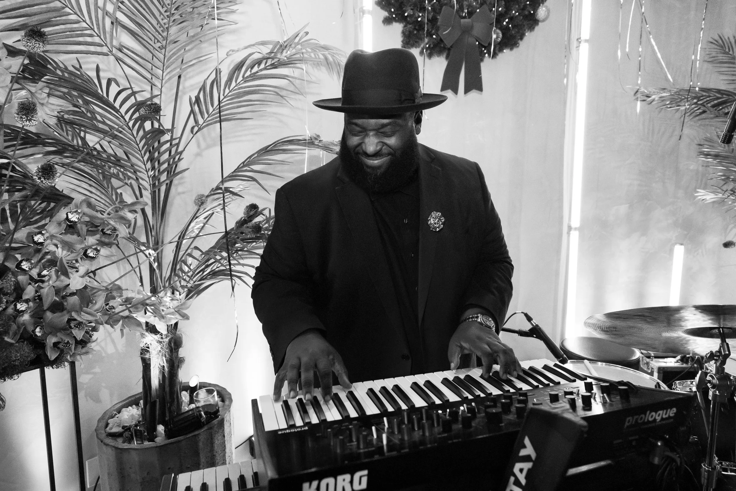 A man wearing a black hat, black suit, and a pin on his lapel, playing a keyboard amidst holiday decorations including a Christmas wreath and flowers, with a drum set nearby.