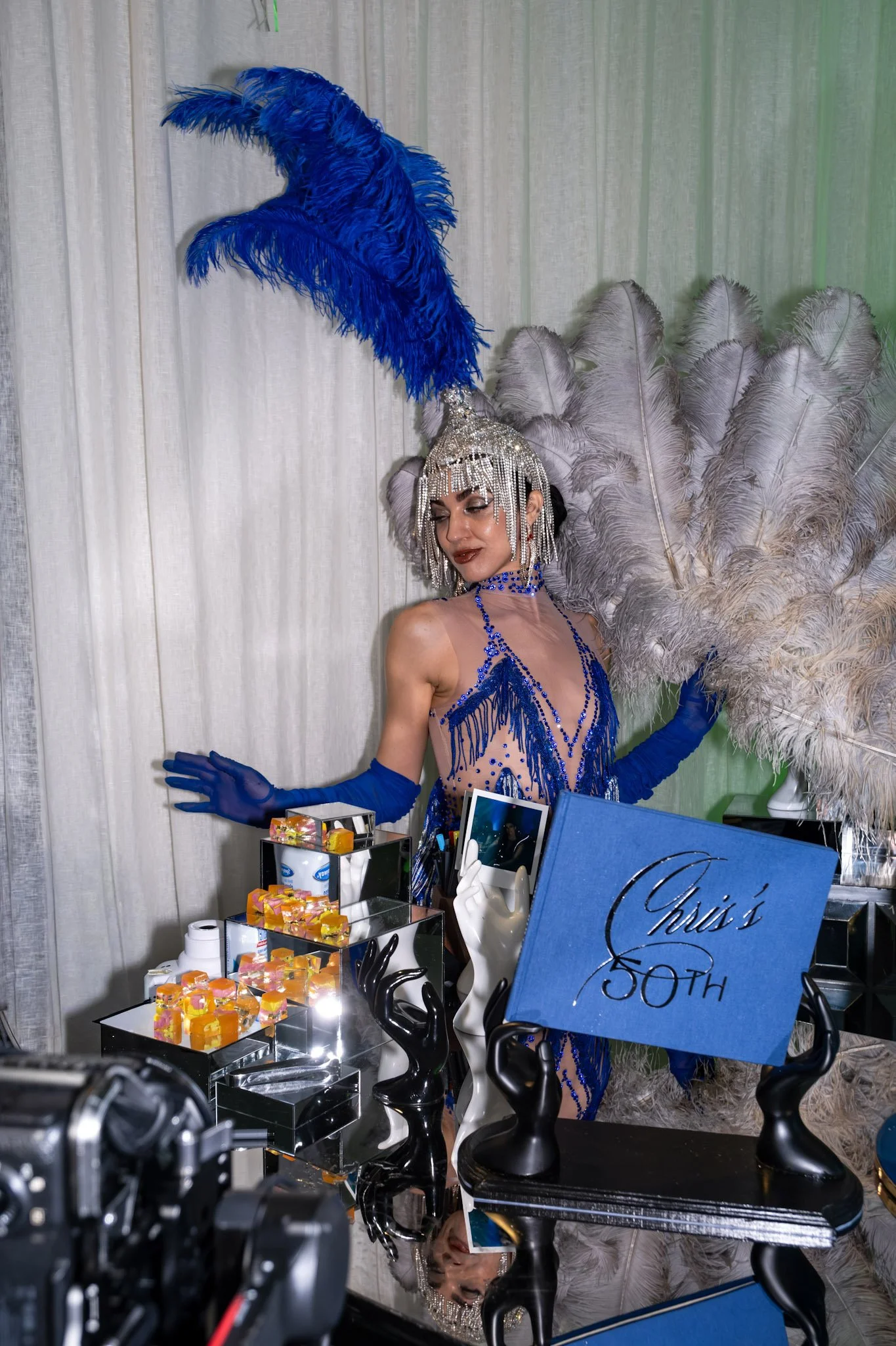 A woman dressed in a blue sheer dress with fringe and beads, wearing long blue gloves, a bejeweled headdress with large blue feathers, and holding white feathers, standing in front of a backdrop of large light-colored feathers. There are various decorative items and a sign that reads 'Christ 50th' on the table in front of her.