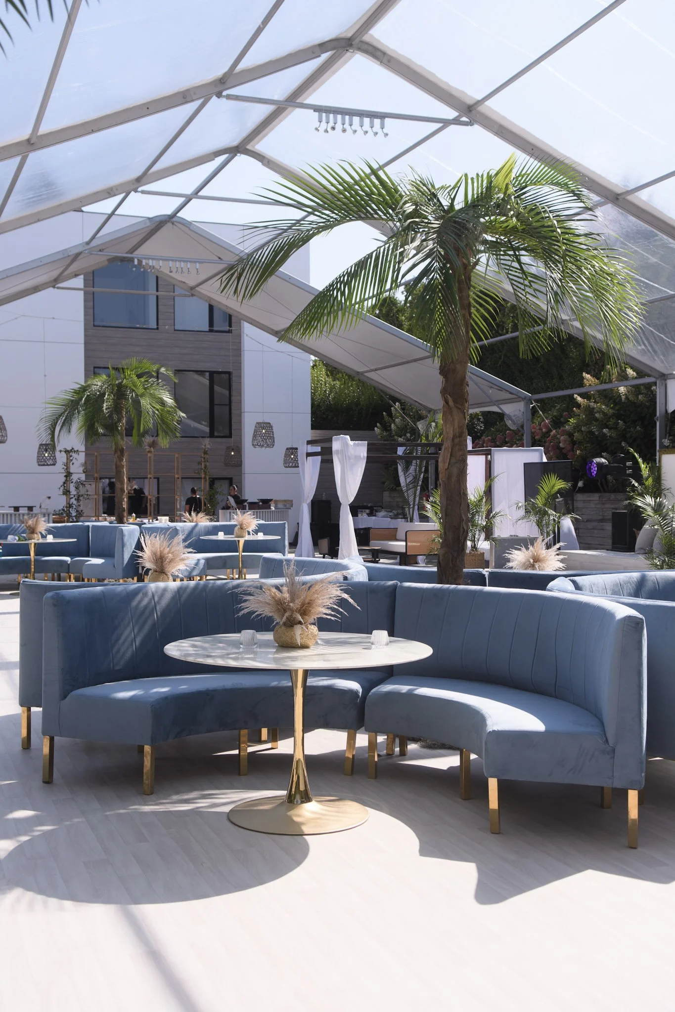 Under a large transparent tent, there are blue velvet sectional sofas, small round tables with decorative vases filled with pampas grass, tall indoor palm trees, and background guests, creating a luxurious event space.