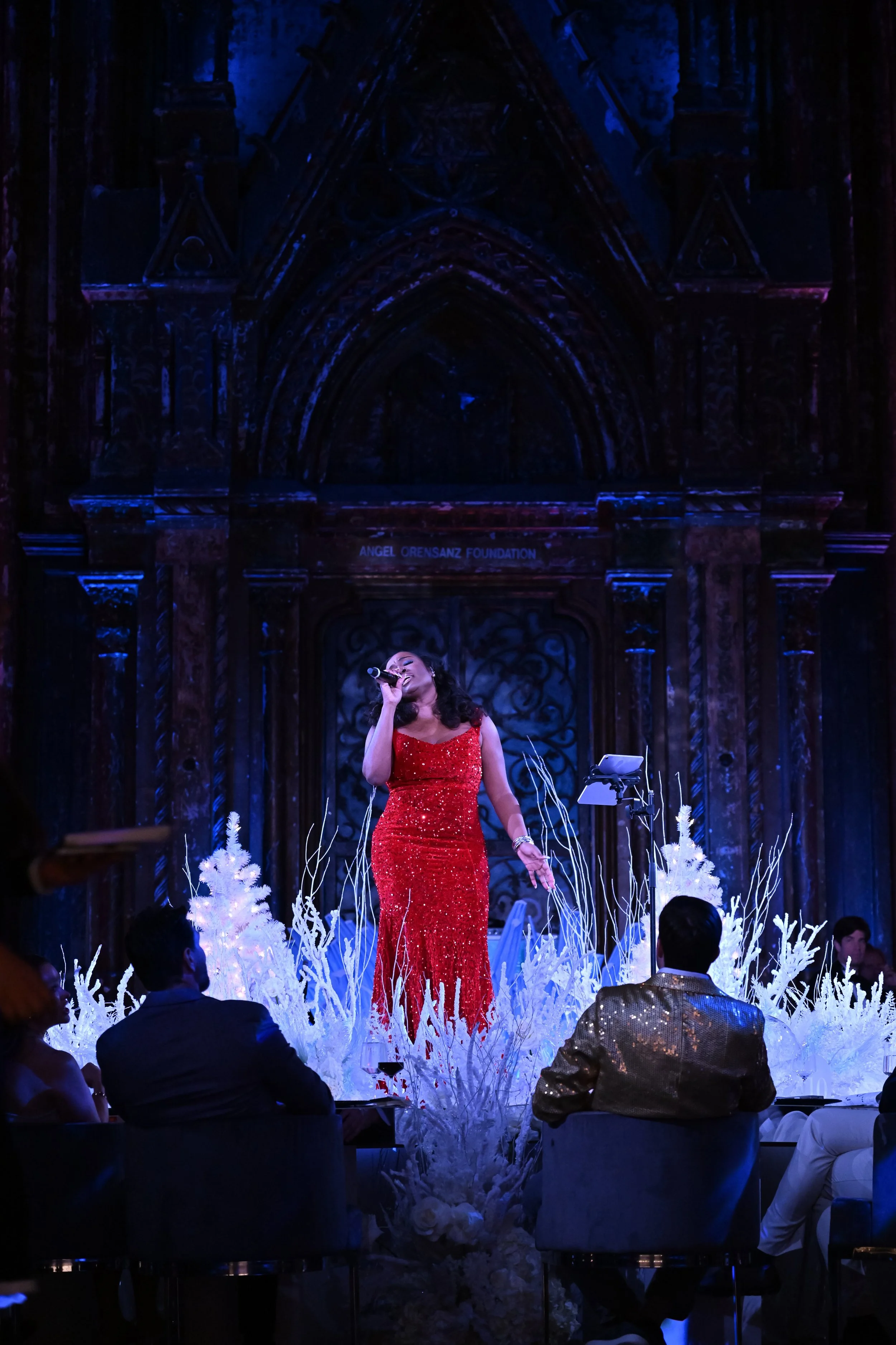 A woman in a sparkling red gown singing into a microphone on a decorated stage with white Christmas trees, surrounded by seated guests.