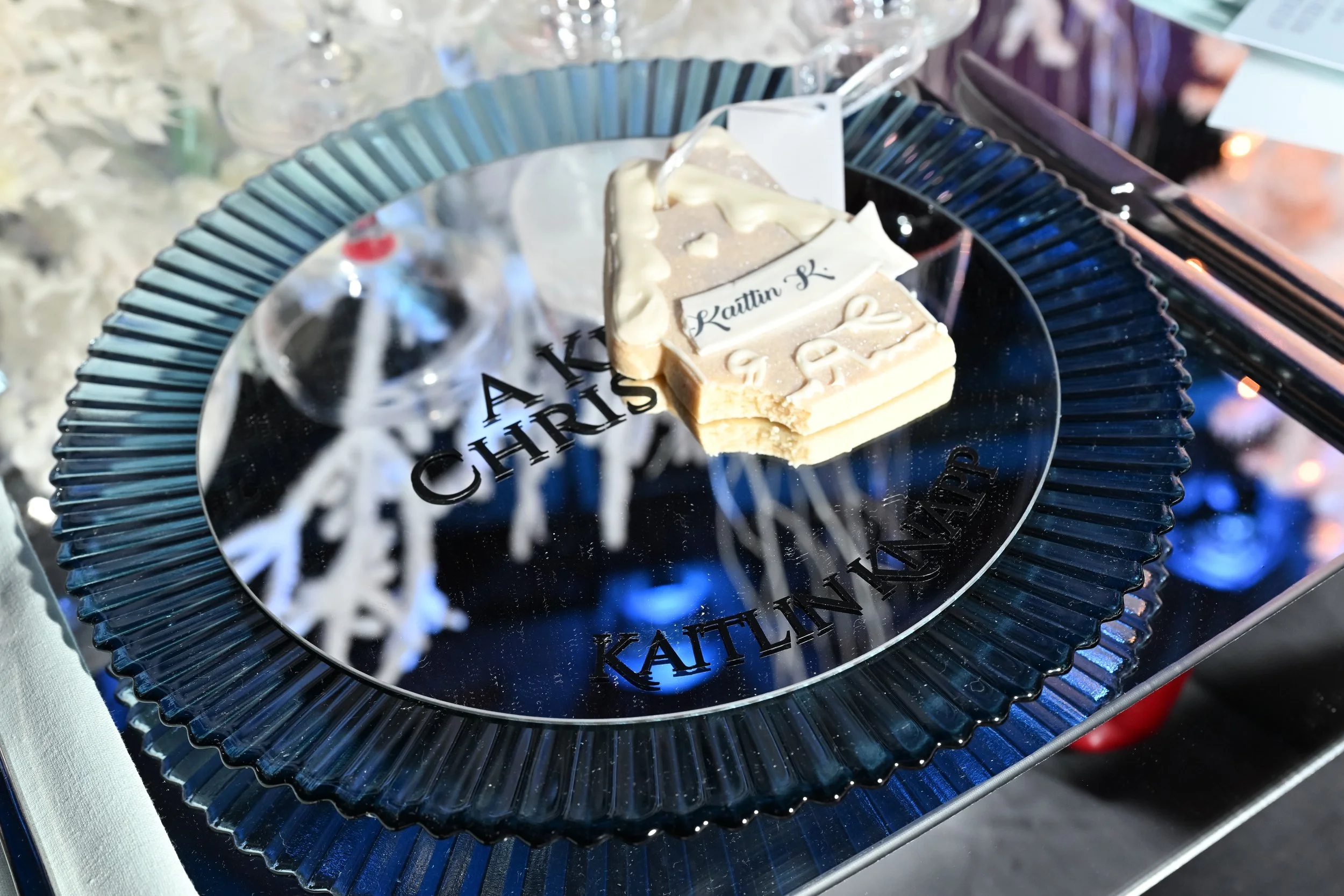 A celebratory cake with white and beige frosting on a reflective silver platter that has black text reading 'ALEX CHRIS' and 'Kaitlin K.' The cake is positioned on a blue glass plate with a fluted edge, set on a table with some wine glasses and decorative elements in the background.