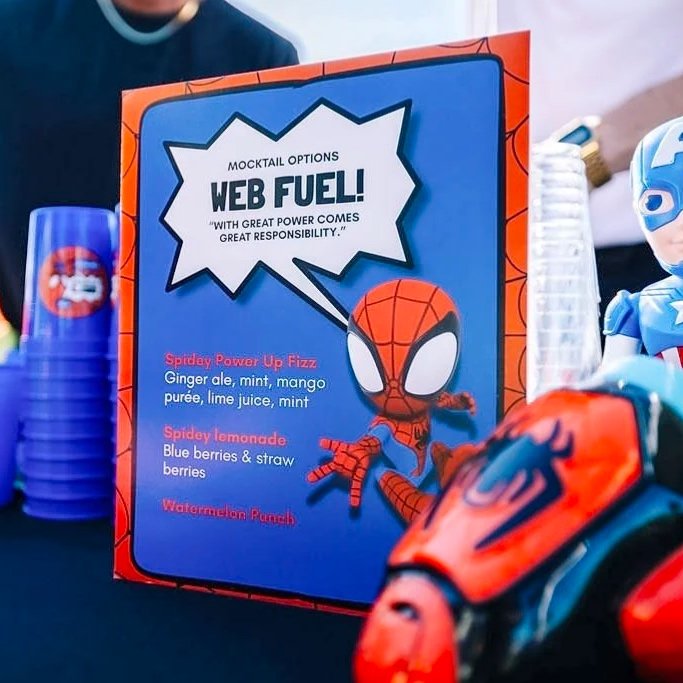 A colorful table display features a Spider-Man themed menu board listing various mocktail options, including Spidey Power Up Fizz, Spidey lemonade, and Watermelon Punch, accompanied by Spider-Man and superhero-themed toys.