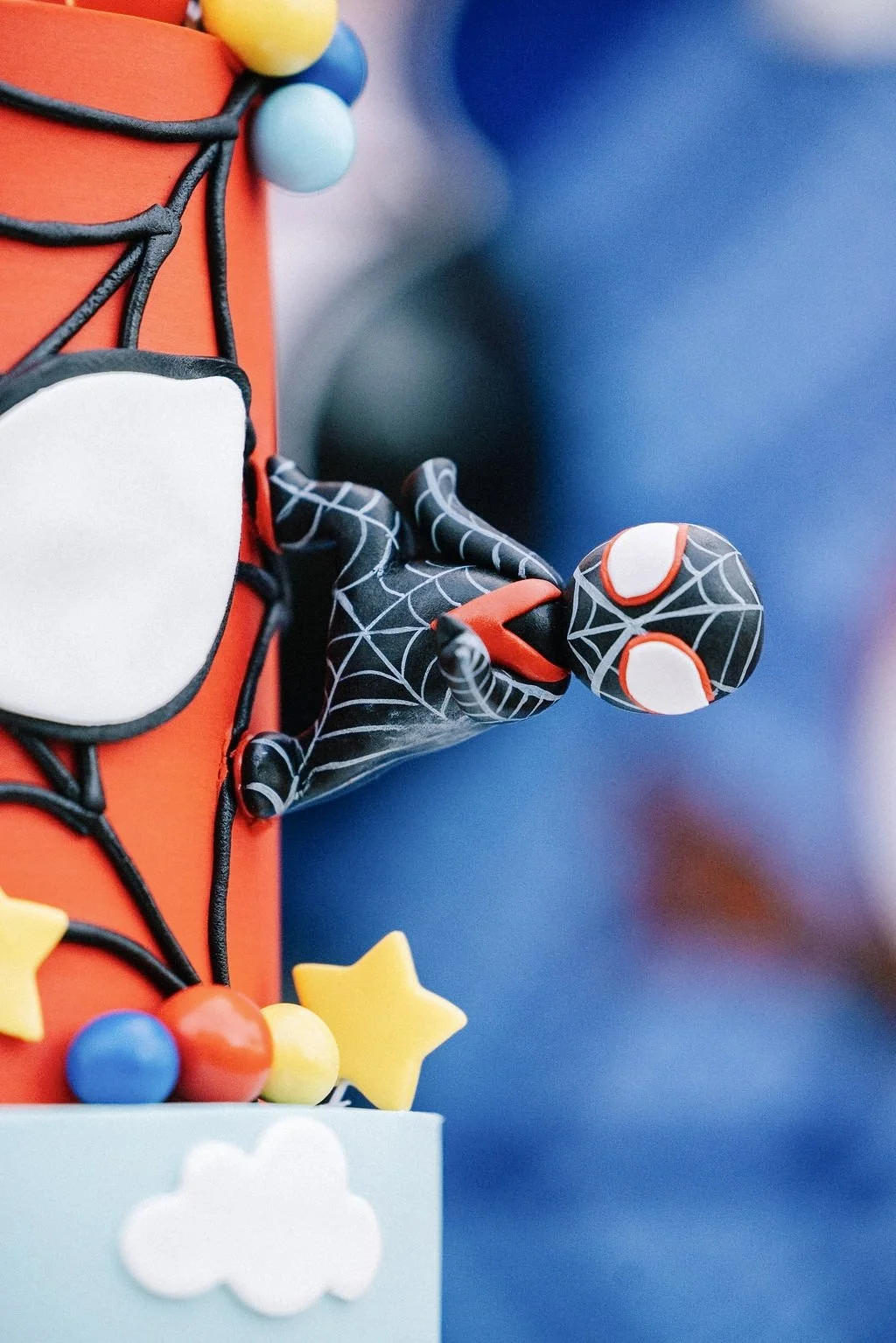 A toy Spider-Man figure is attached to a vibrant birthday cake decorated with colorful candies, stars, and cloud shapes.