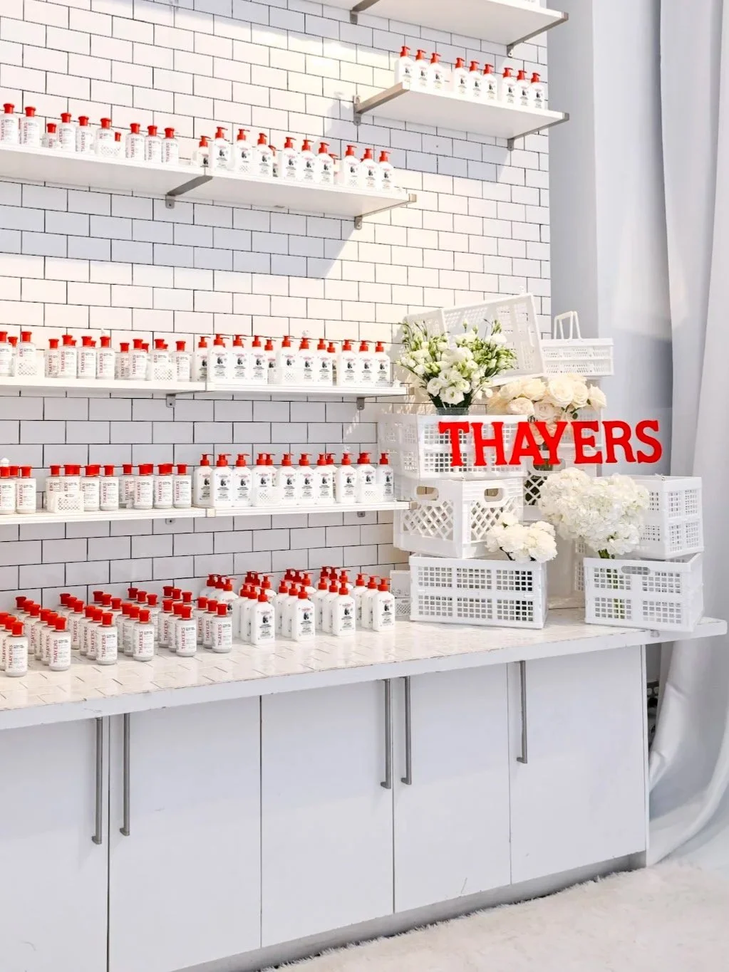 Display of white bottles with red caps on white shelves against a white subway tile wall, with floral arrangements and a red sign that reads "THAYERS".