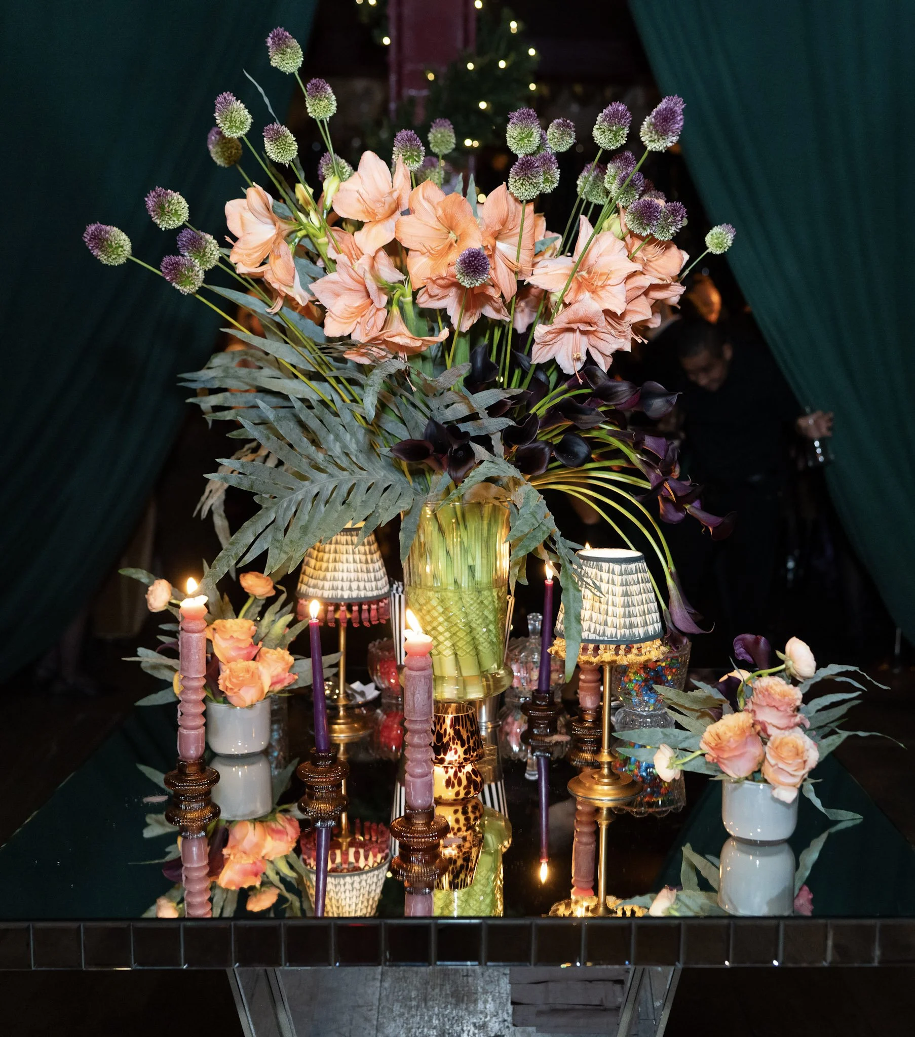 Elegant floral centerpiece with orange and purple flowers, candles, and decorative lamps on a reflective table at an indoor event.