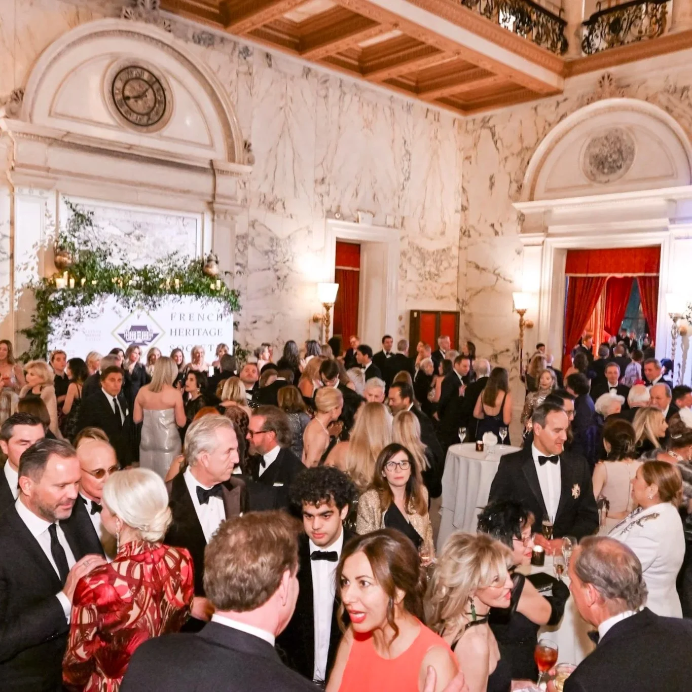 Guests dressed in formal attire at a crowded reception in an elegant hall with marble walls, high ceilings, tall windows with red curtains, and a decorated archway.