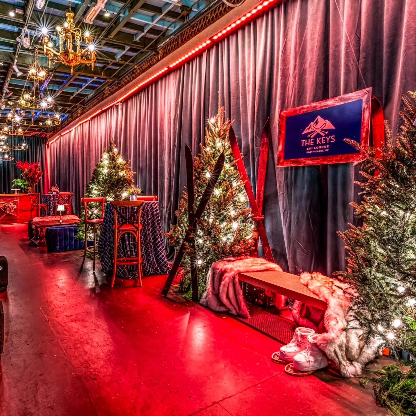 Indoor scene at a ski lounge decorated for Christmas with Christmas trees, skis, and furry accessories. There are tables and chairs, a large curtain, and a TV screen displaying the logo for The Keys Ski Lounge in East Village, NY.