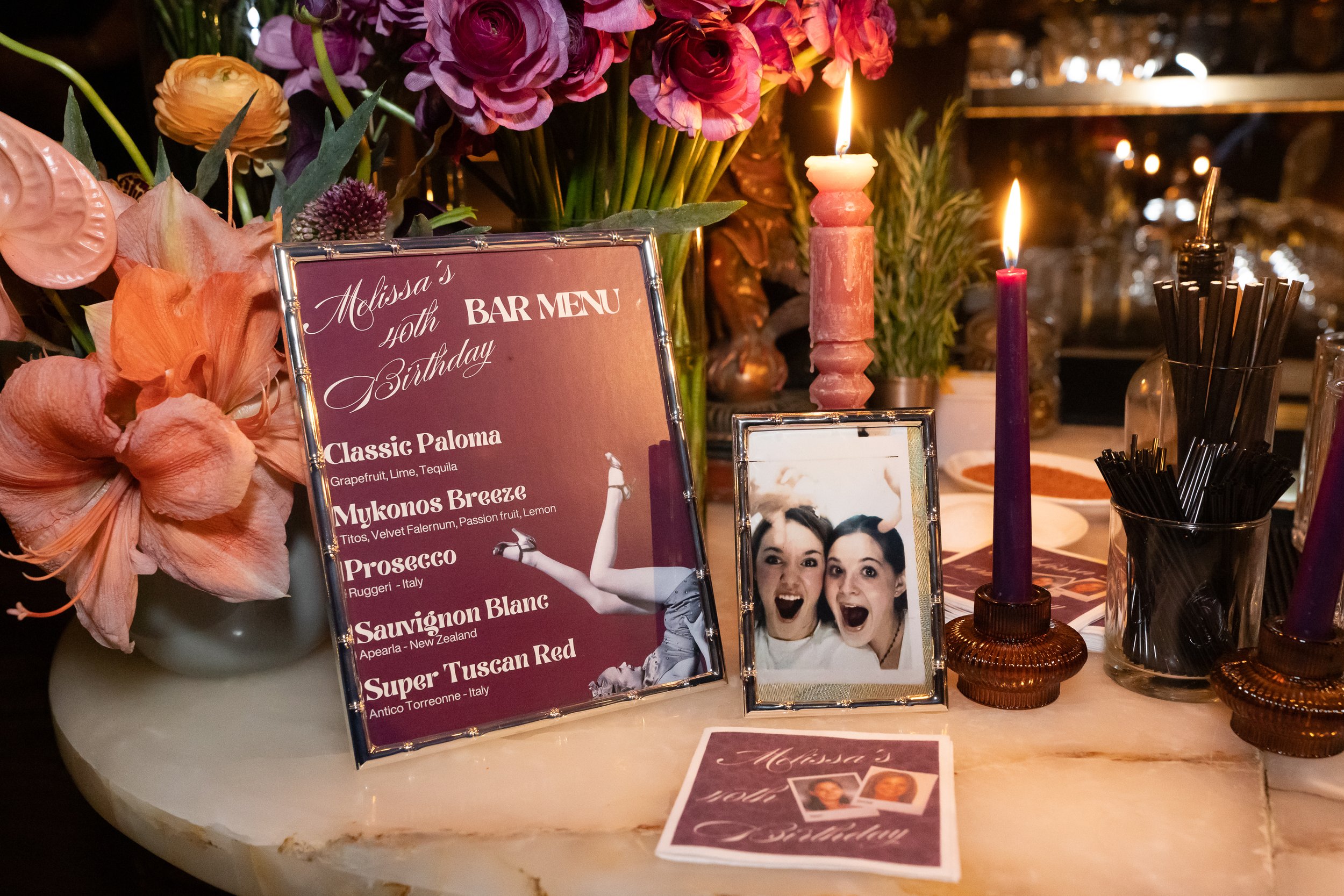 Decorative table with pink flowers, lit candles, a framed photo of two women, a bar menu sign, and a small birthday card, celebrating Melissa's 40th birthday.