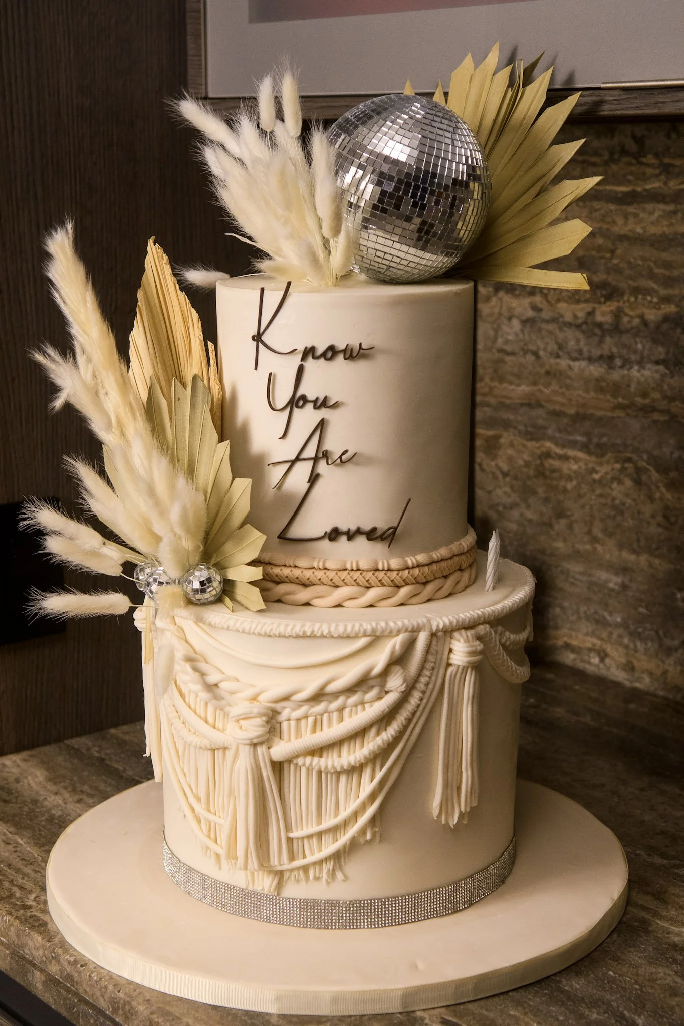 A two-tiered cream-colored cake with decorative draping and tassels, topped with a disco ball, pampas grass, and metallic leaves, with the phrase 'Know You Are Loved' written on the top tier.
