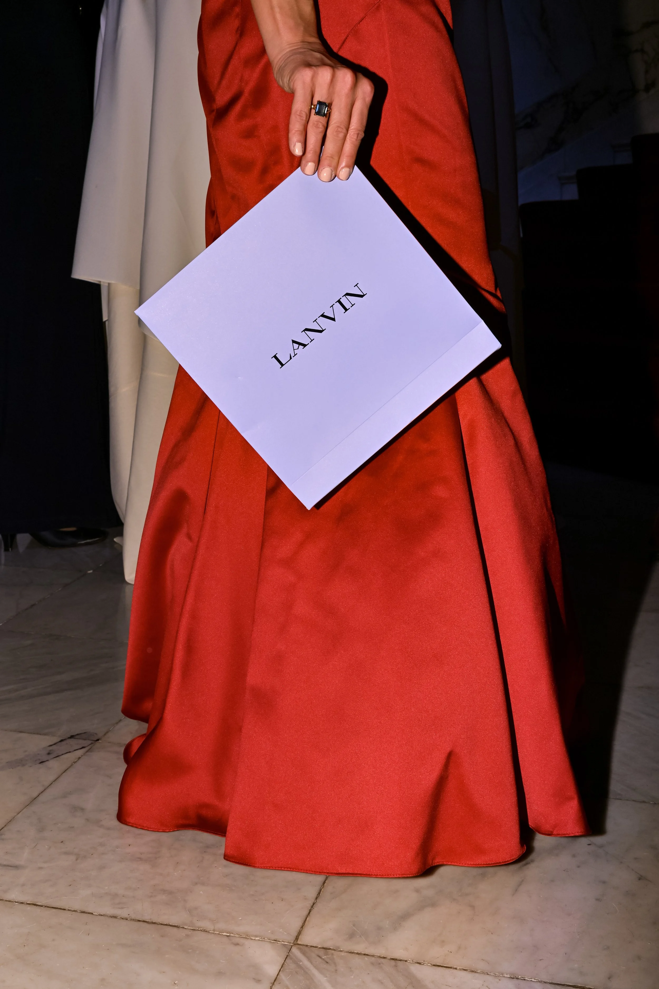 Close-up of a person holding a Lanvin shopping bag, wearing a red satin dress and a ring, with other garments hanging in the background.