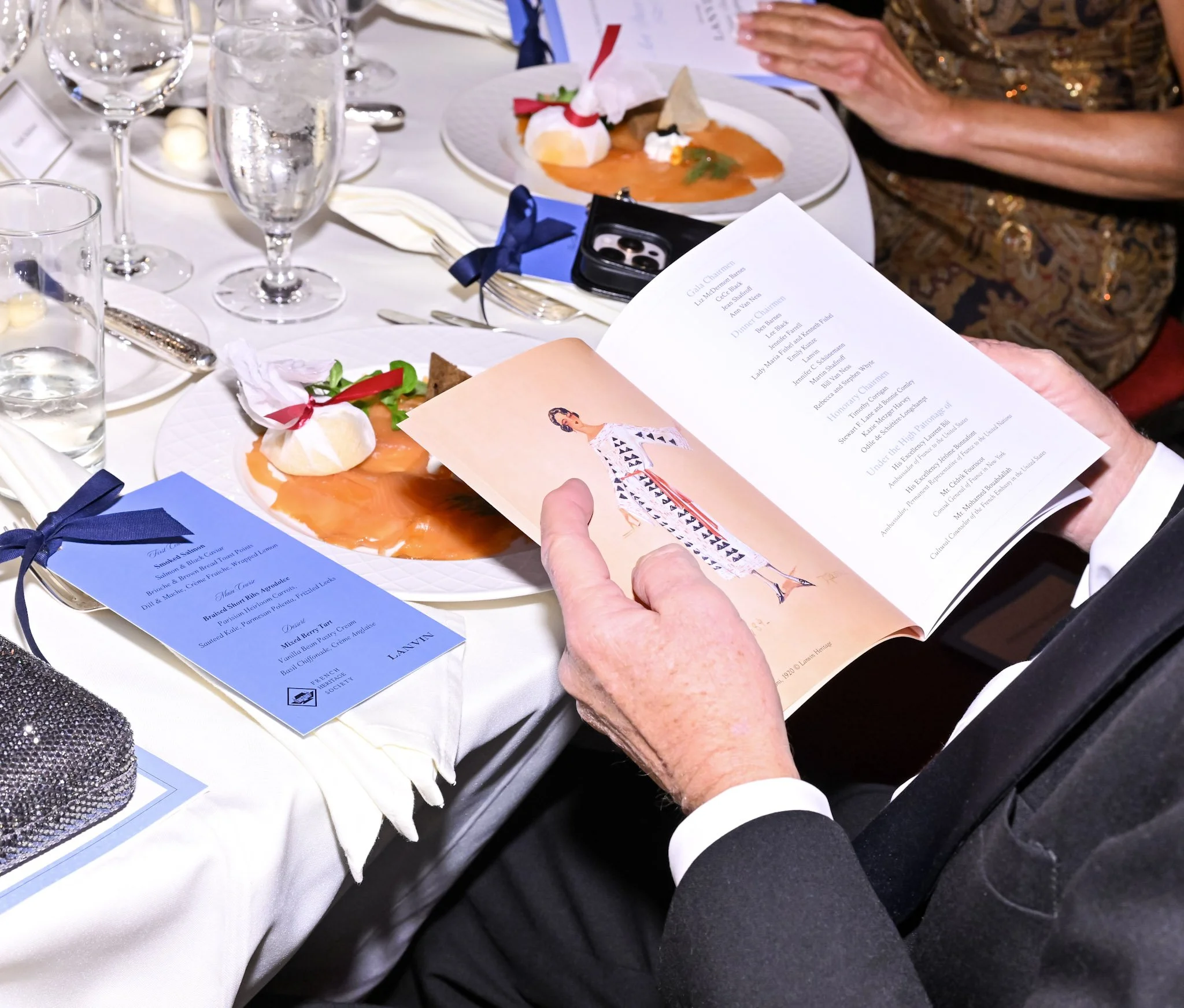 Person in a tuxedo reading a program at a formal dinner table with plates of food, glasses of water, and blue menus.