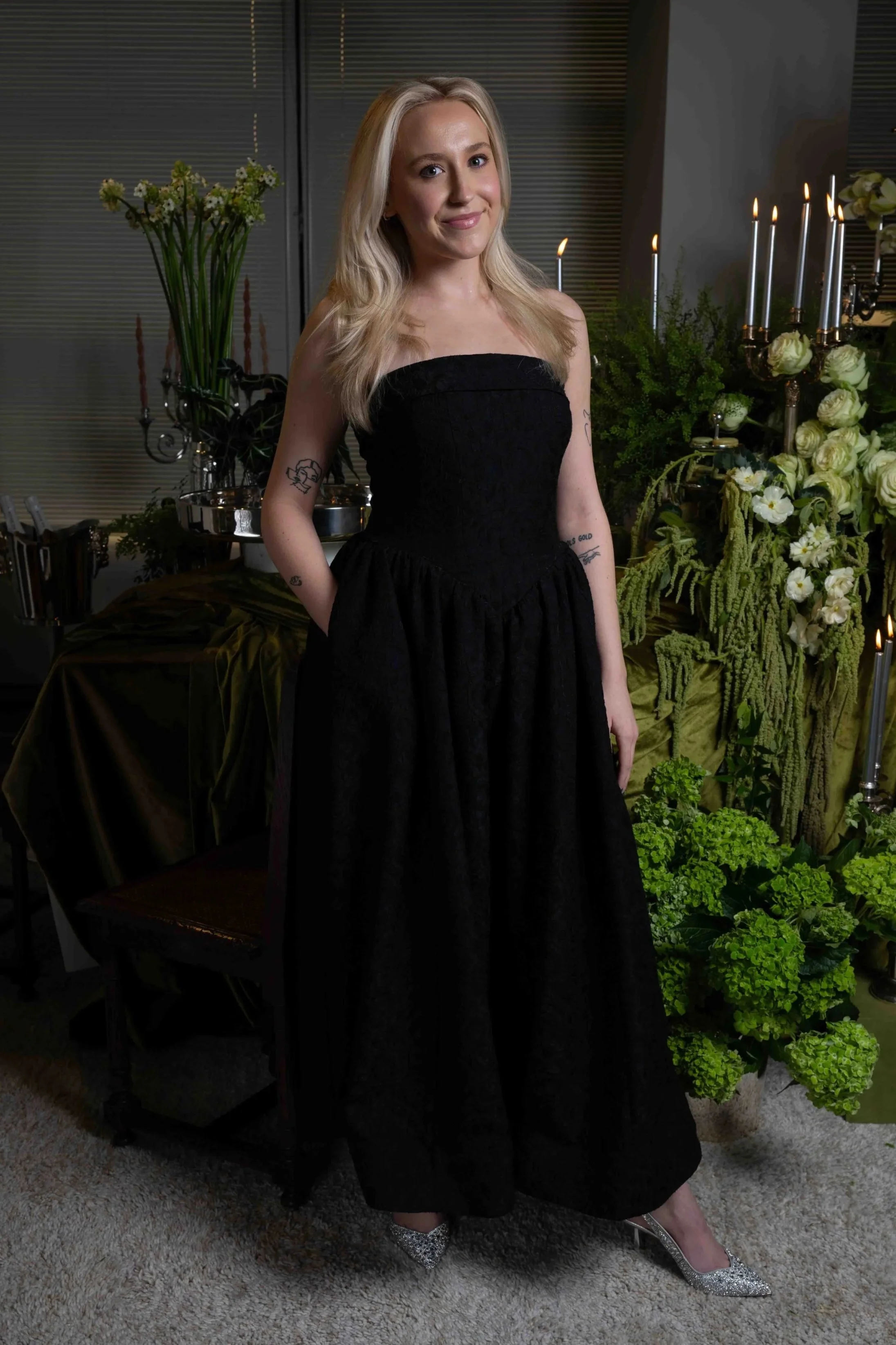 A woman in a strapless black dress and silver heels standing indoors near a table with green plants and white flowers, and lit candles in the background.