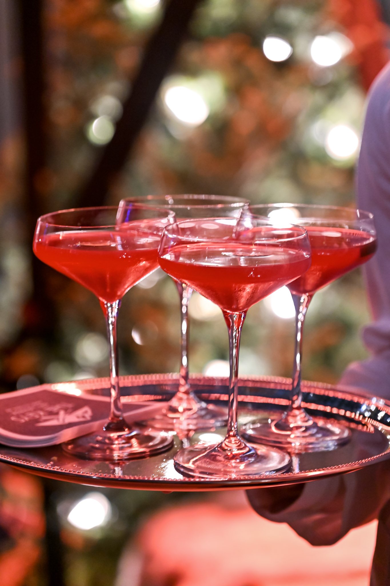 Four coupe glasses filled with red wine on a red tray, held by a person wearing a light-colored shirt, with a colorful blurred outdoor background.
