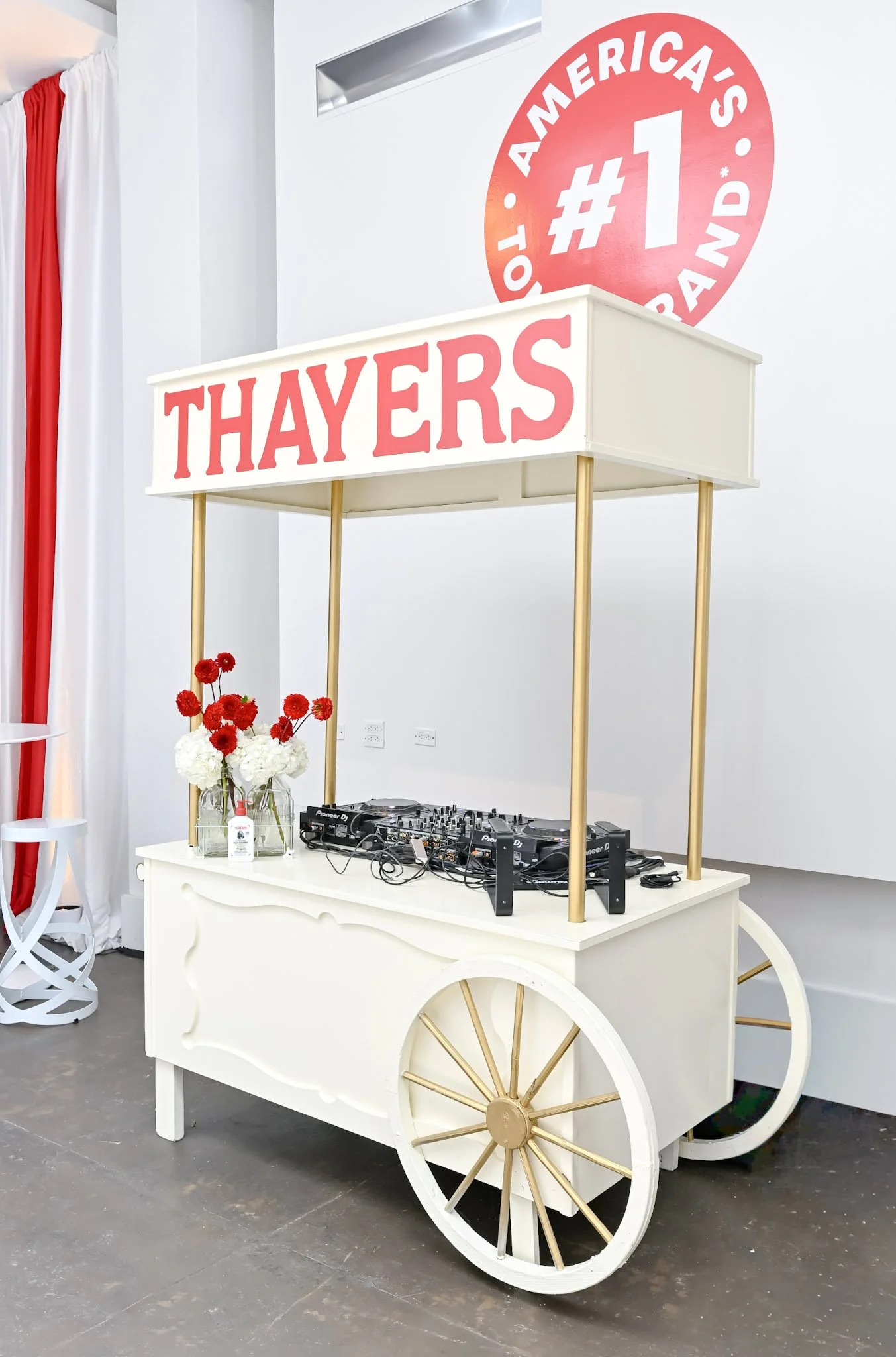 A white cart with gold accents and wheels, serving as a DJ booth with mixing equipment and flowers, under a sign that reads 'Thayers' and a backdrop with 'America's #1'