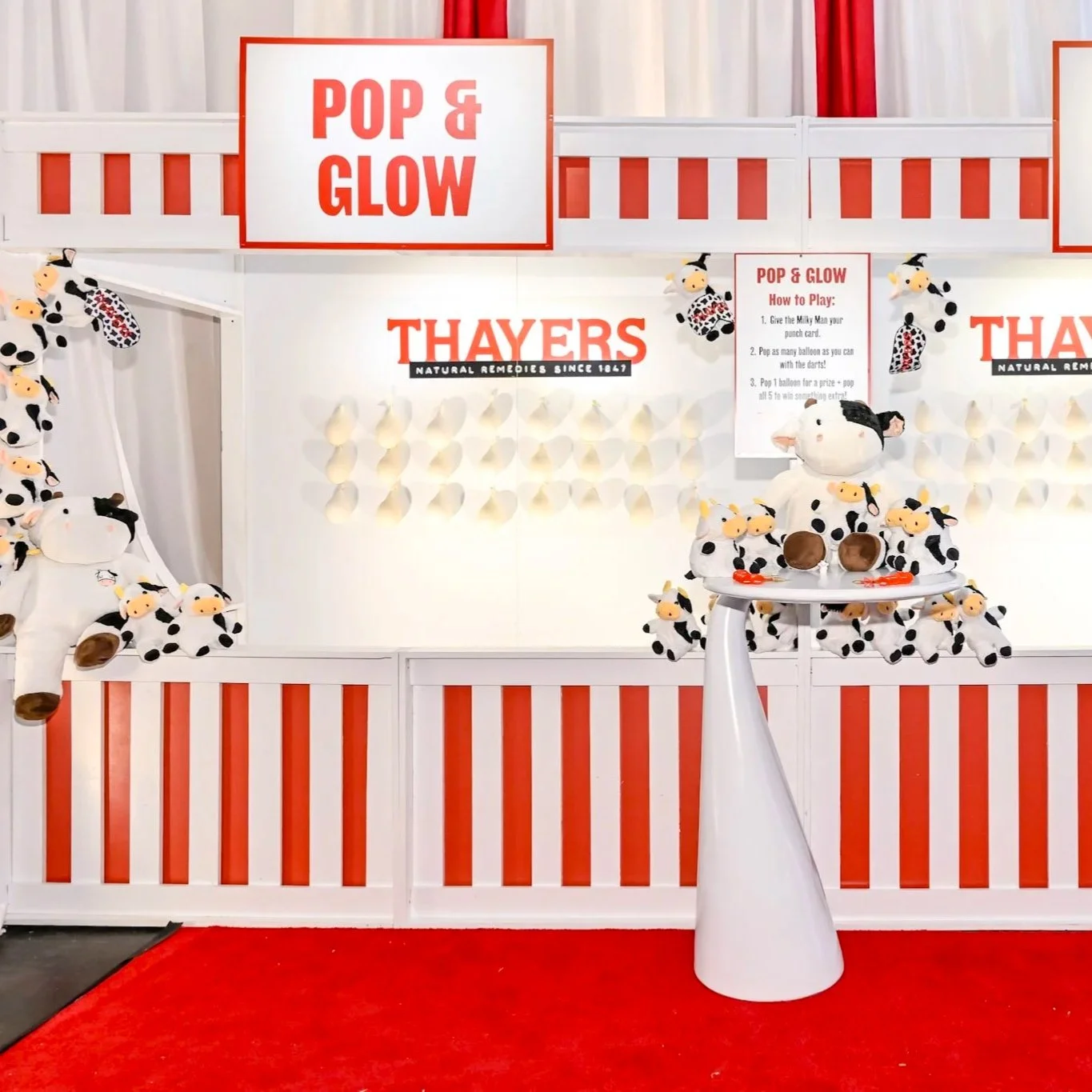 Display featuring plush cow toys and a large plush cow, with a red and white theme, promotional signs, and instructions for a game called 'Pop & Glow' at Thayers, a natural remedies company.