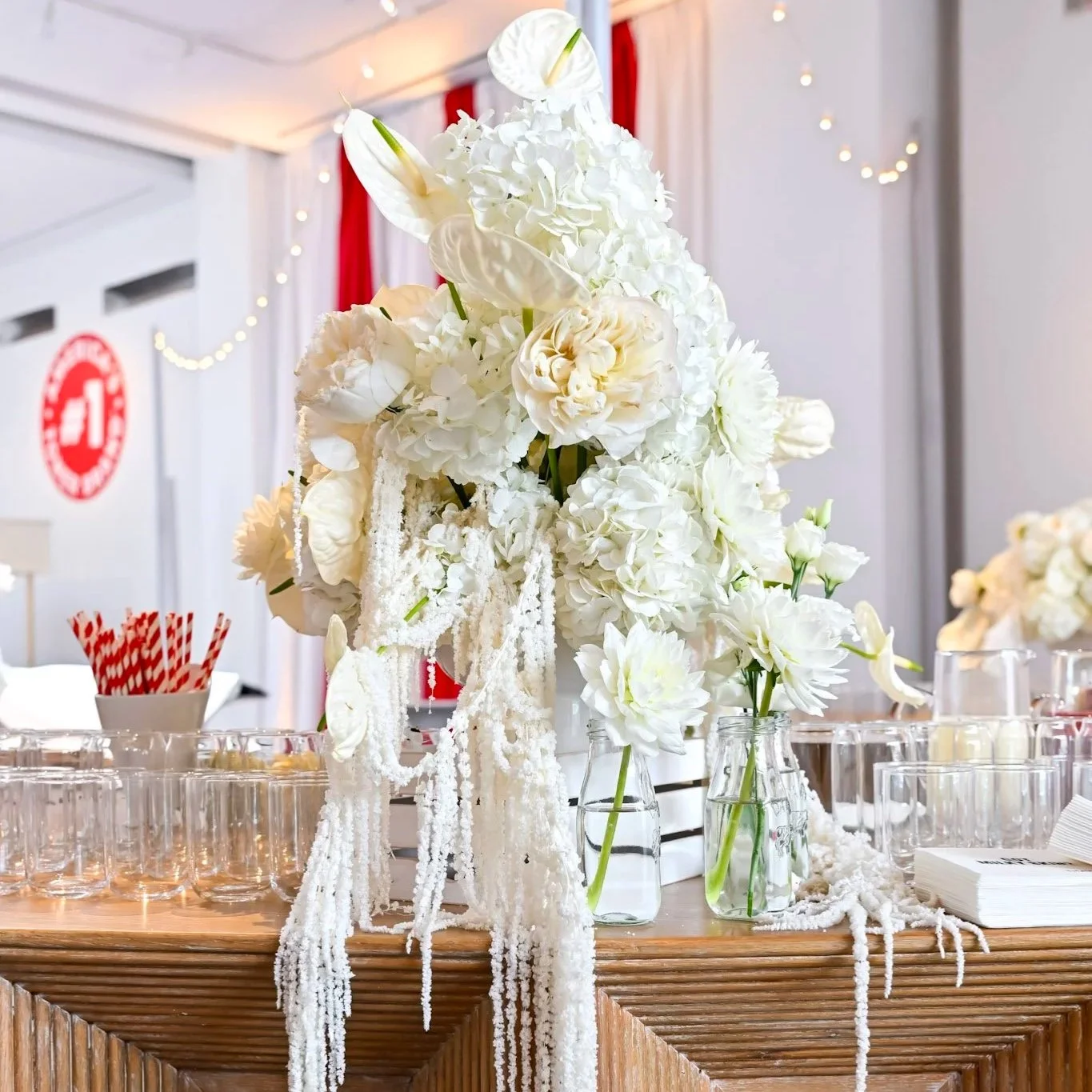 White floral centerpiece with roses, hydrangeas, and calla lilies on a wooden table, decorated with white draping and string lights in the background.