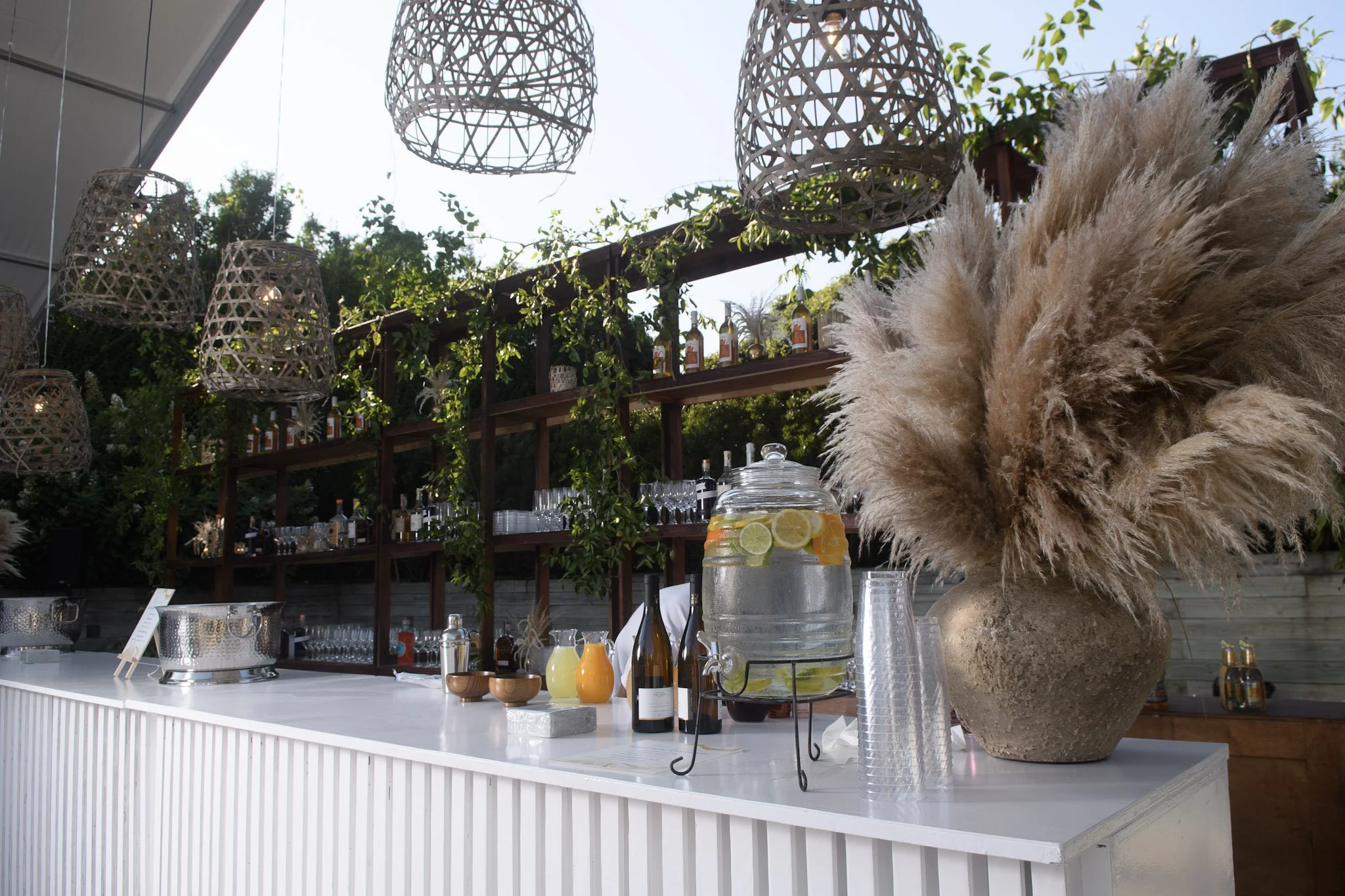 Outdoor bar set up with a large vase of pampas grass, a glass jar of lemon and lime infused water, bottles of alcohol, and various drinks on a white counter. Hanging wicker lanterns and greenery decorate the background.