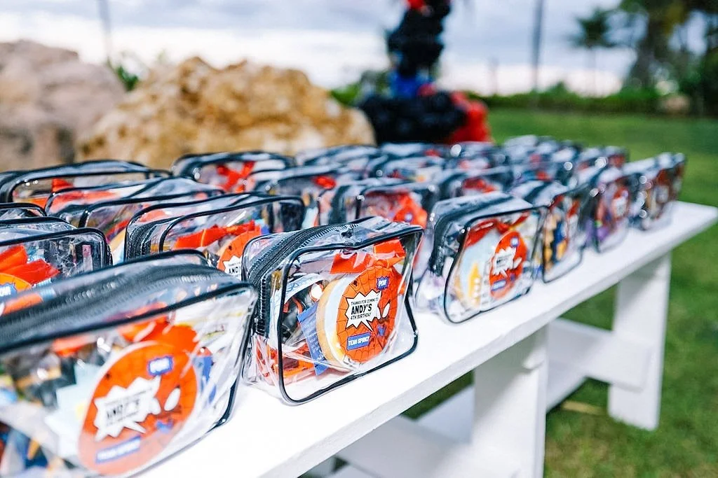 Clear plastic tote bags with orange, white, and blue labels, lined up on a white picnic table outdoors.