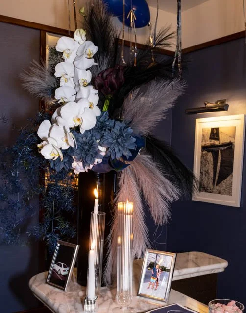A floral arrangement with white orchids, pink and blue flowers, and dark feathers, displayed on a table with lit candles, framed photos, and artwork in the background.
