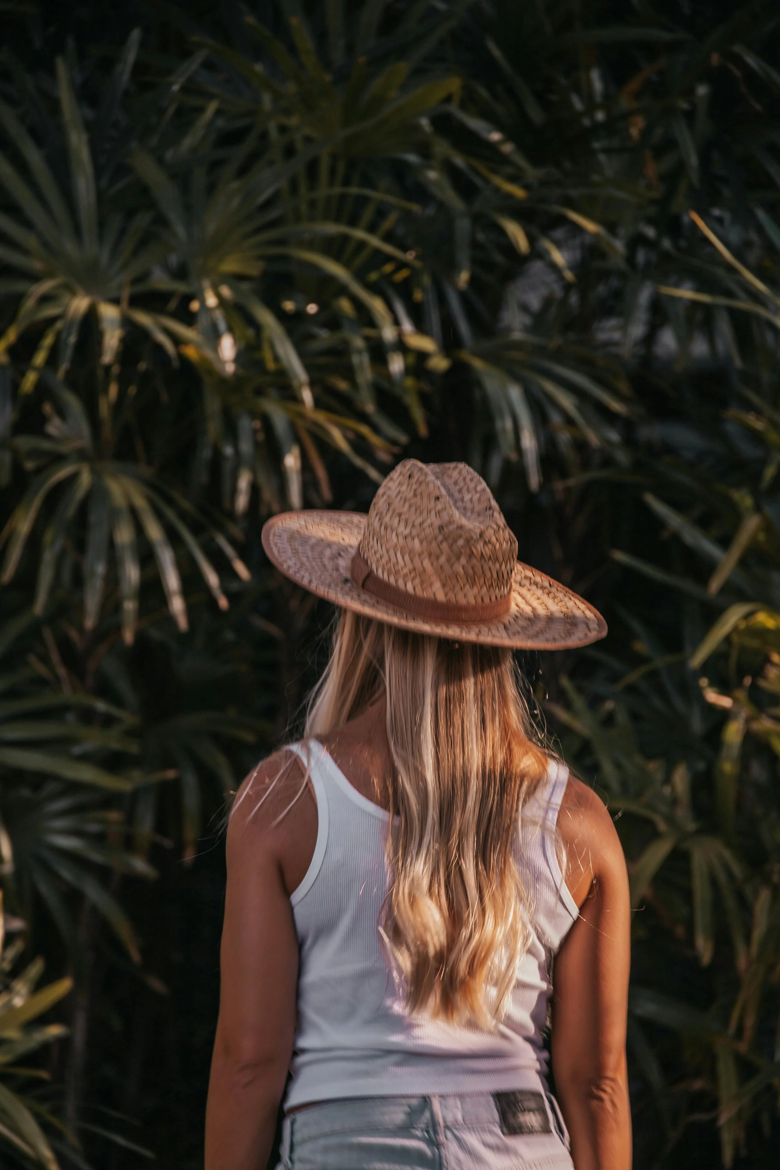 A woman with long blonde hair wearing a white tank top and a straw hat, standing in front of lush green tropical plants. Healthy blonde hair with natural layers, inspired by the effortless styles offered at Cosmic Beauty Kapolei.