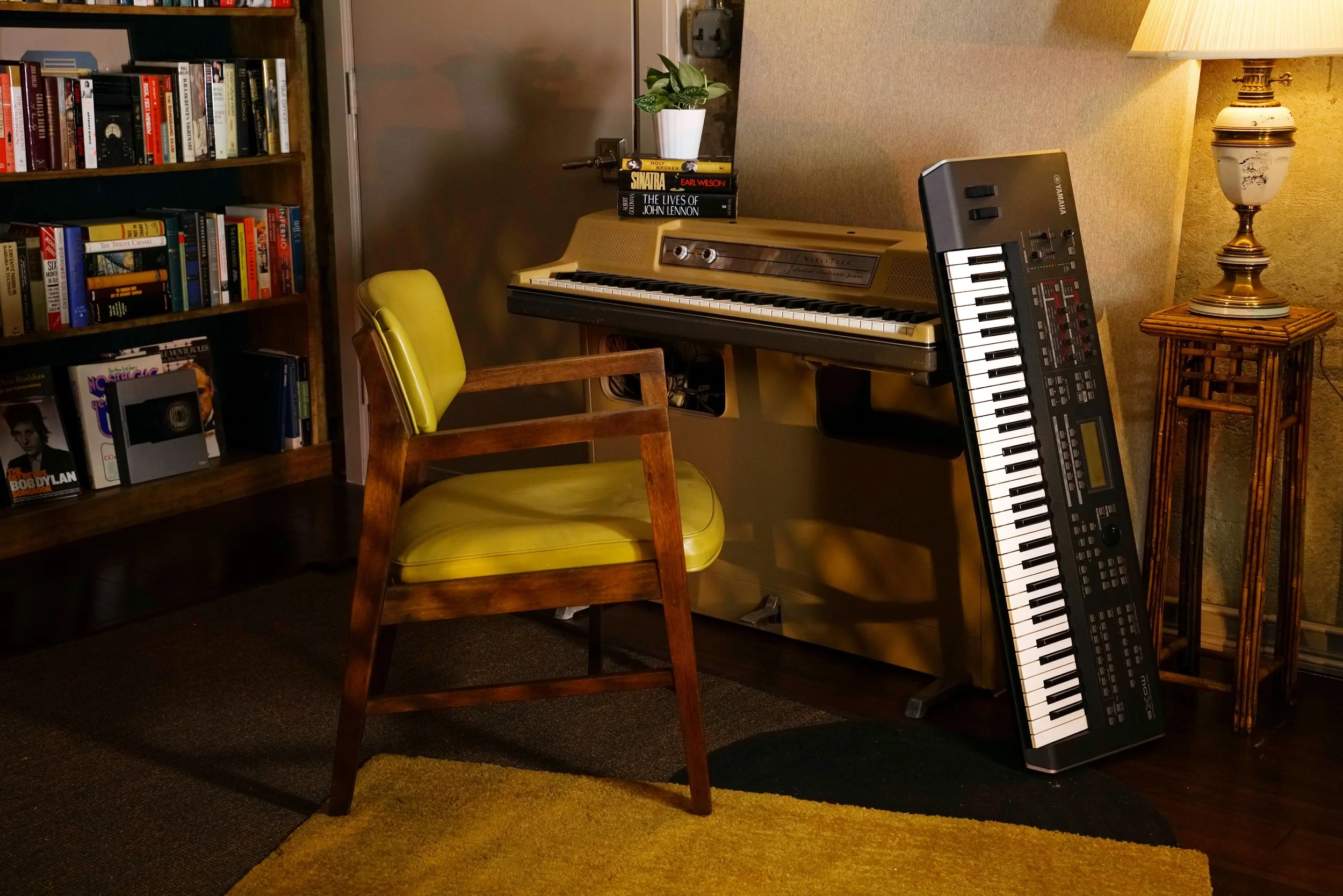 A Wurlitzer 206 with a yellow chair positioned so that someone could walk up and play, and a Yamaha workstation propped up against the electronic piano.