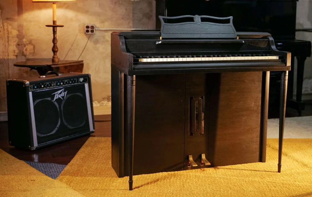 A Wurlitzer 700 electronic piano, refinished in black, in a room with concrete walls and yellow carpets. There is a Peavy guitar amplifier at the keyboard's left.