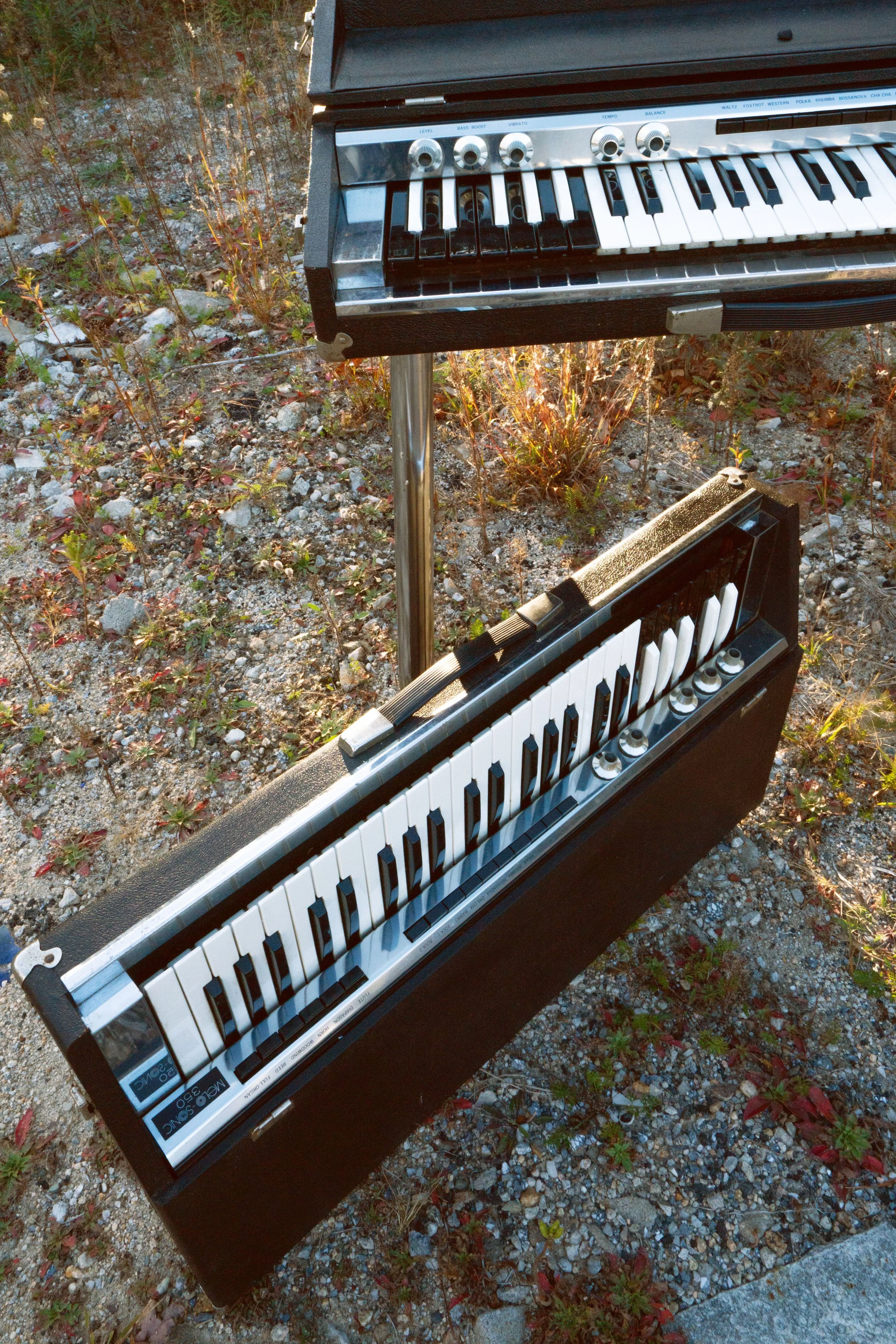 Two Melosonic combo organs, one on chrome legs and one on the ground, in a rocky field.