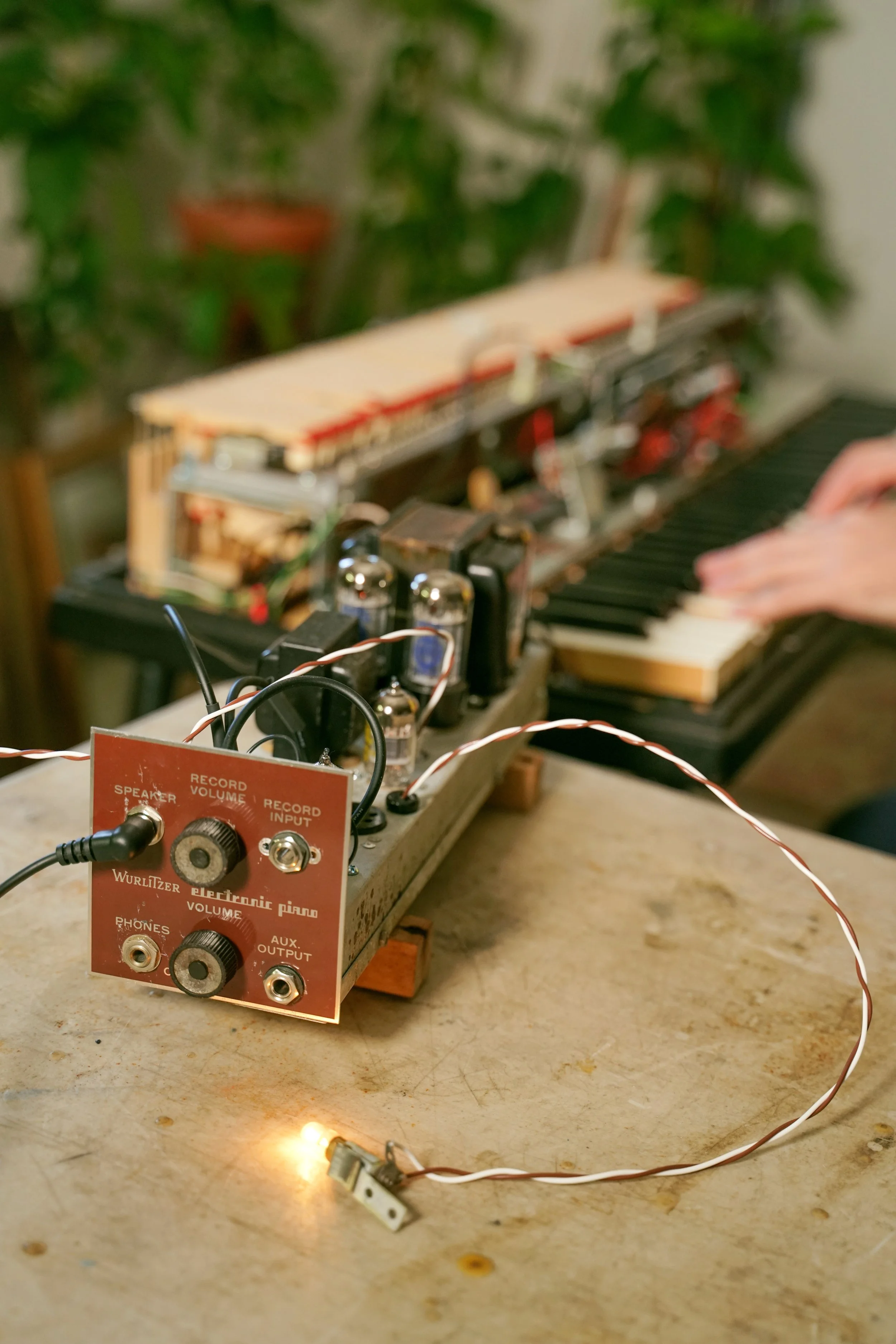 A Wurlitzer 112 amplifier on a table, and a man playing a Wurlitzer 200A in the background.