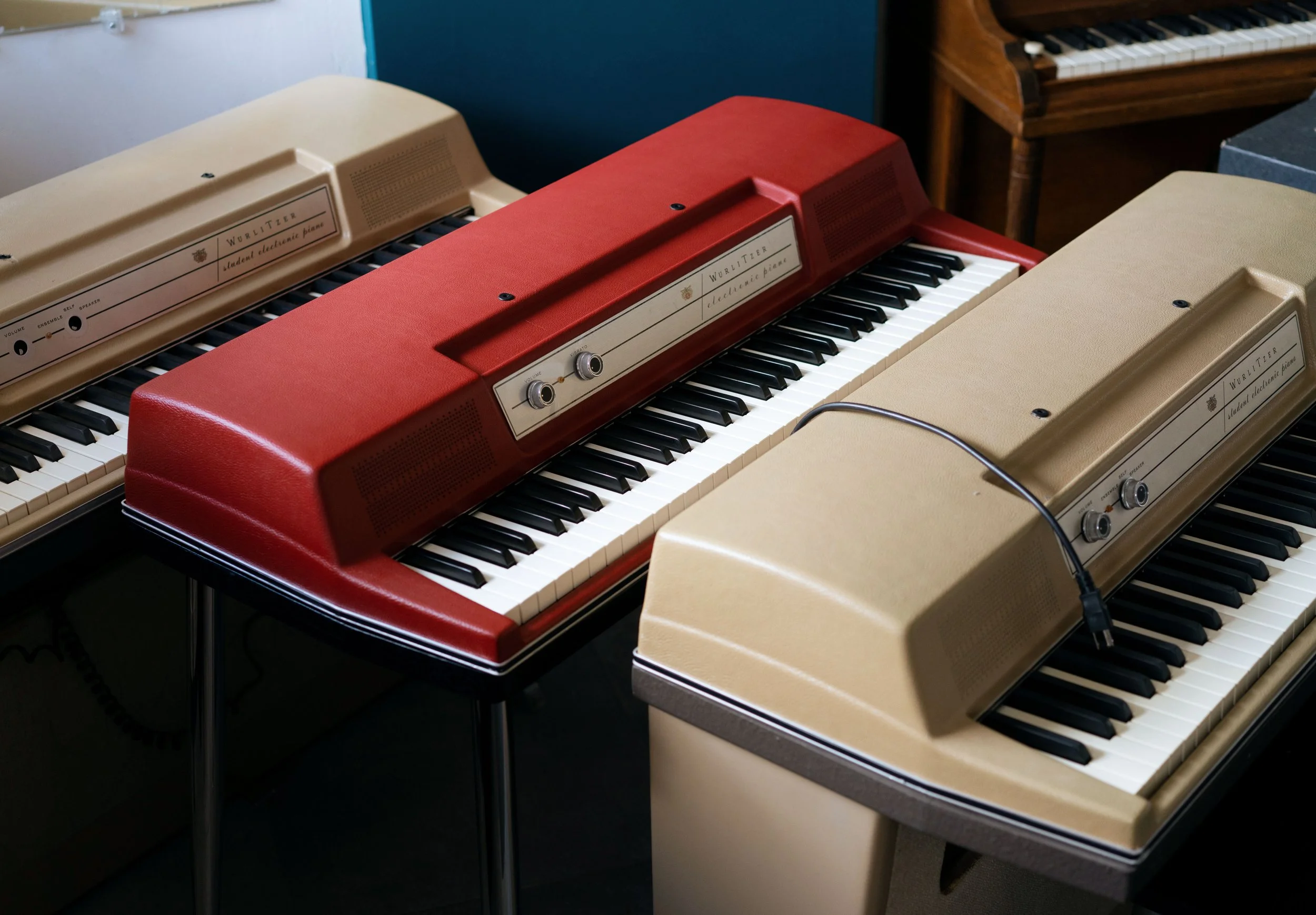 A red Wurlitzer electronic piano pictured between two student-model beige keyboards.