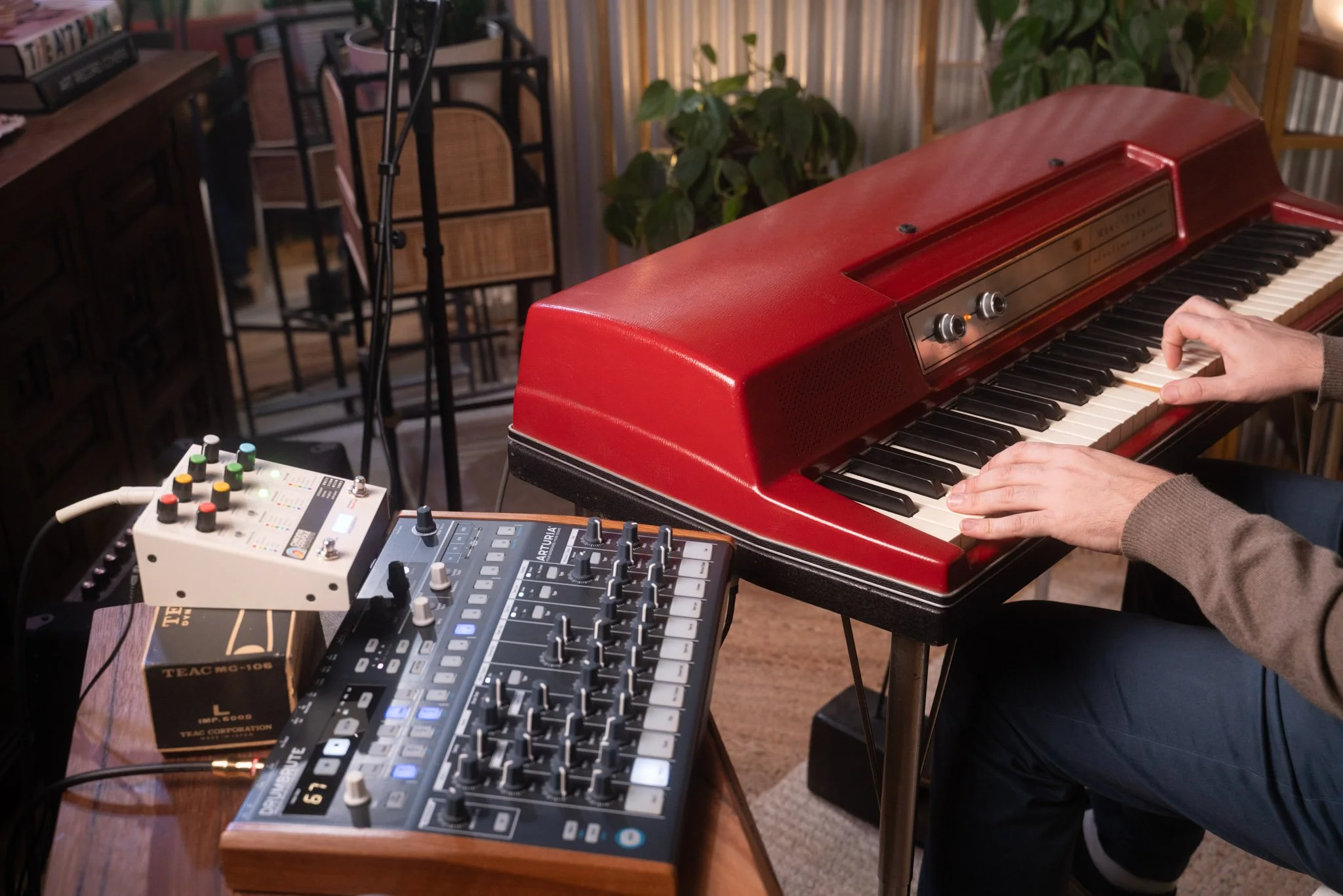 A man playing a Wurlitzer 200 with a red lid, using an effects pedal and a drum machine.