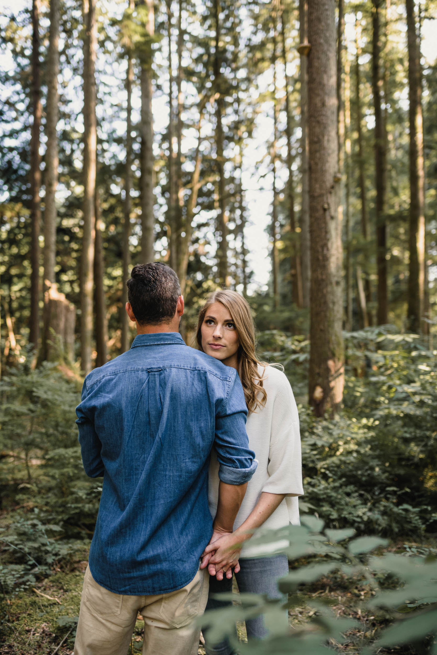 Couple embraces in foliage. Girl looks over guy's shoulder.