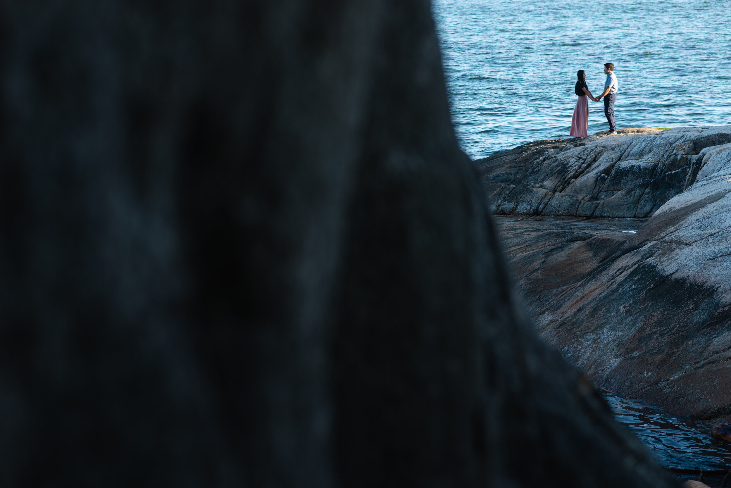 Couple stands on rock next to ocean in distance