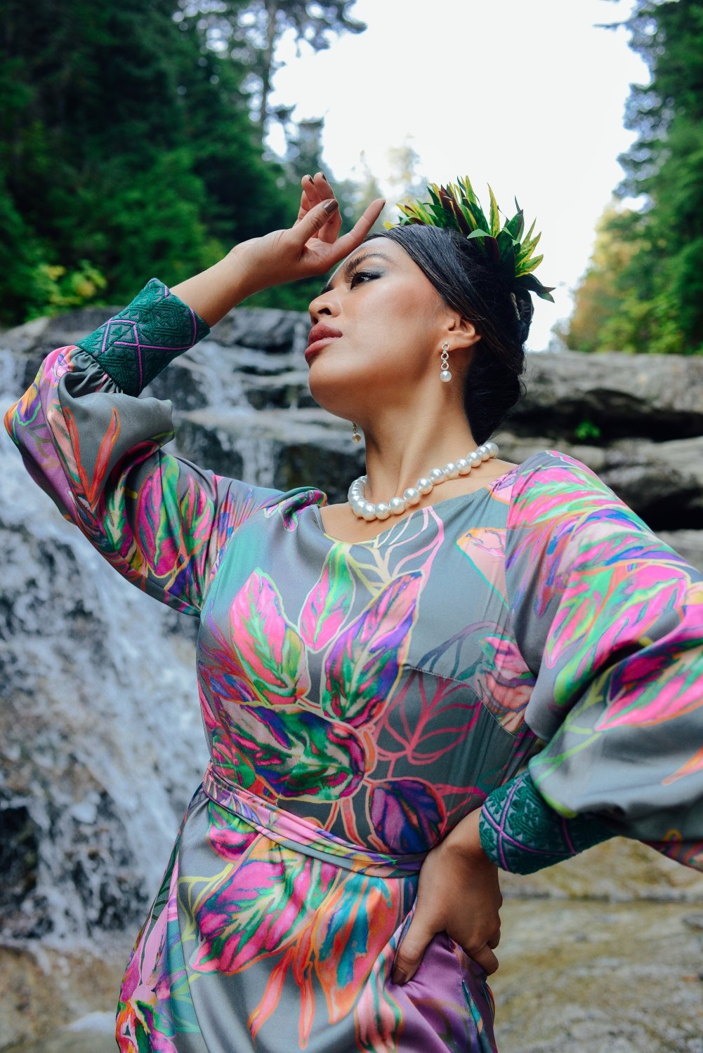 Woman wears a colorful silk gown and stands in the woods at the edge of a waterfall. Pearl jewelry necklace and pearl earrings. Local businesses, female owned. Model dark hair, aapi pacific islander