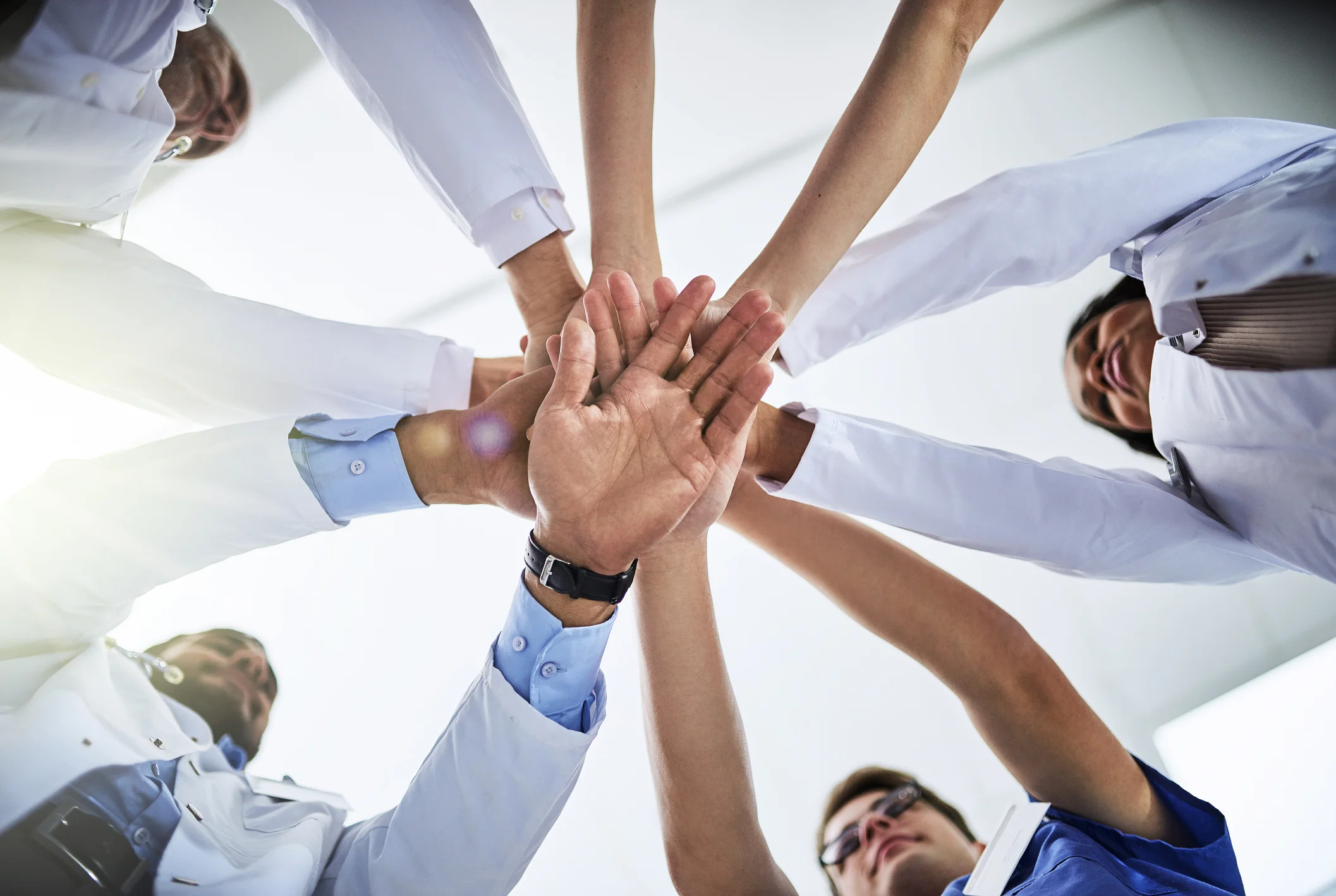 Group of healthcare professionals in medical scrubs and white coats putting their hands together in a teamwork gesture, seen from below.