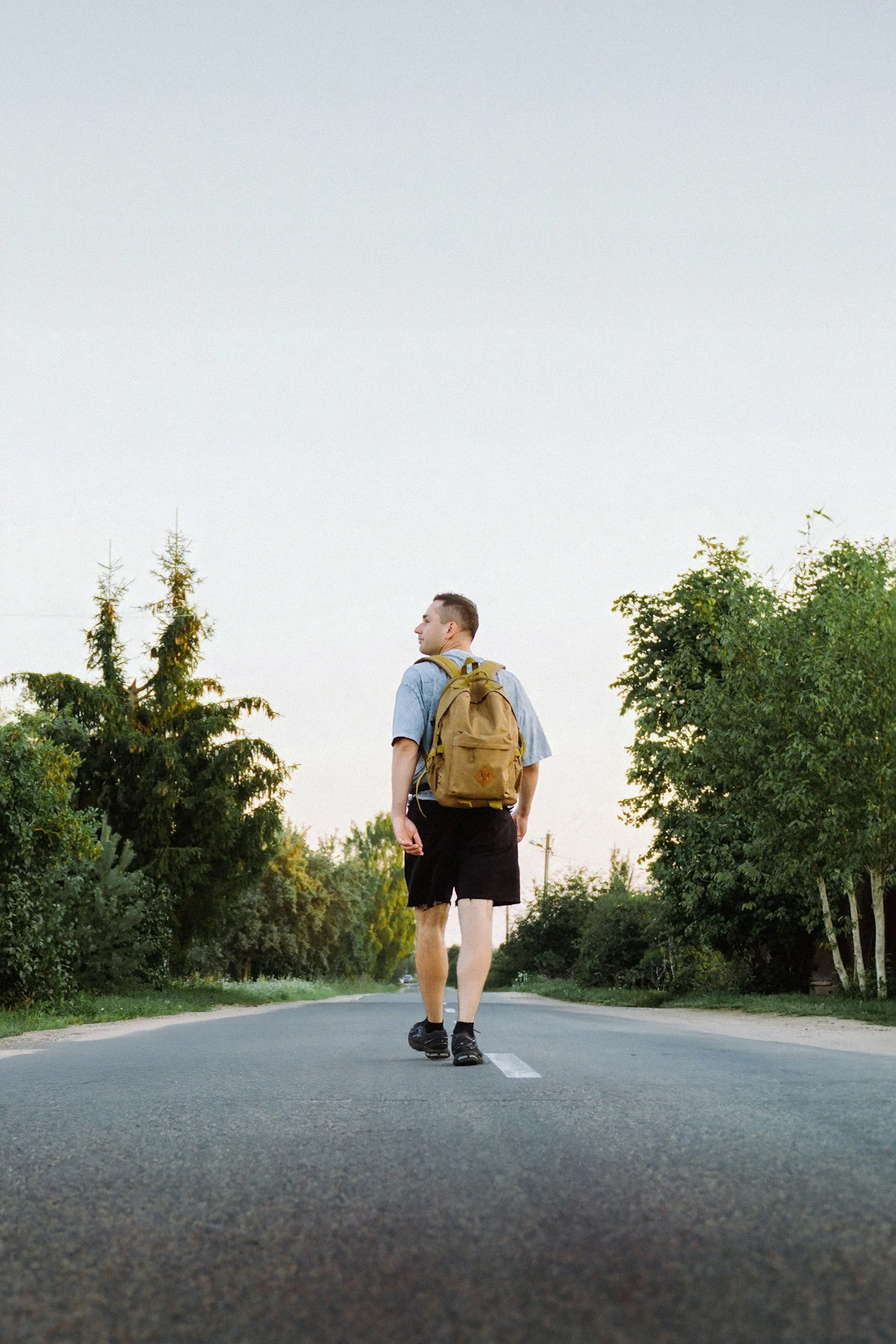 A man with a backpack walking on an empty road during sunset or sunrise, surrounded by trees.