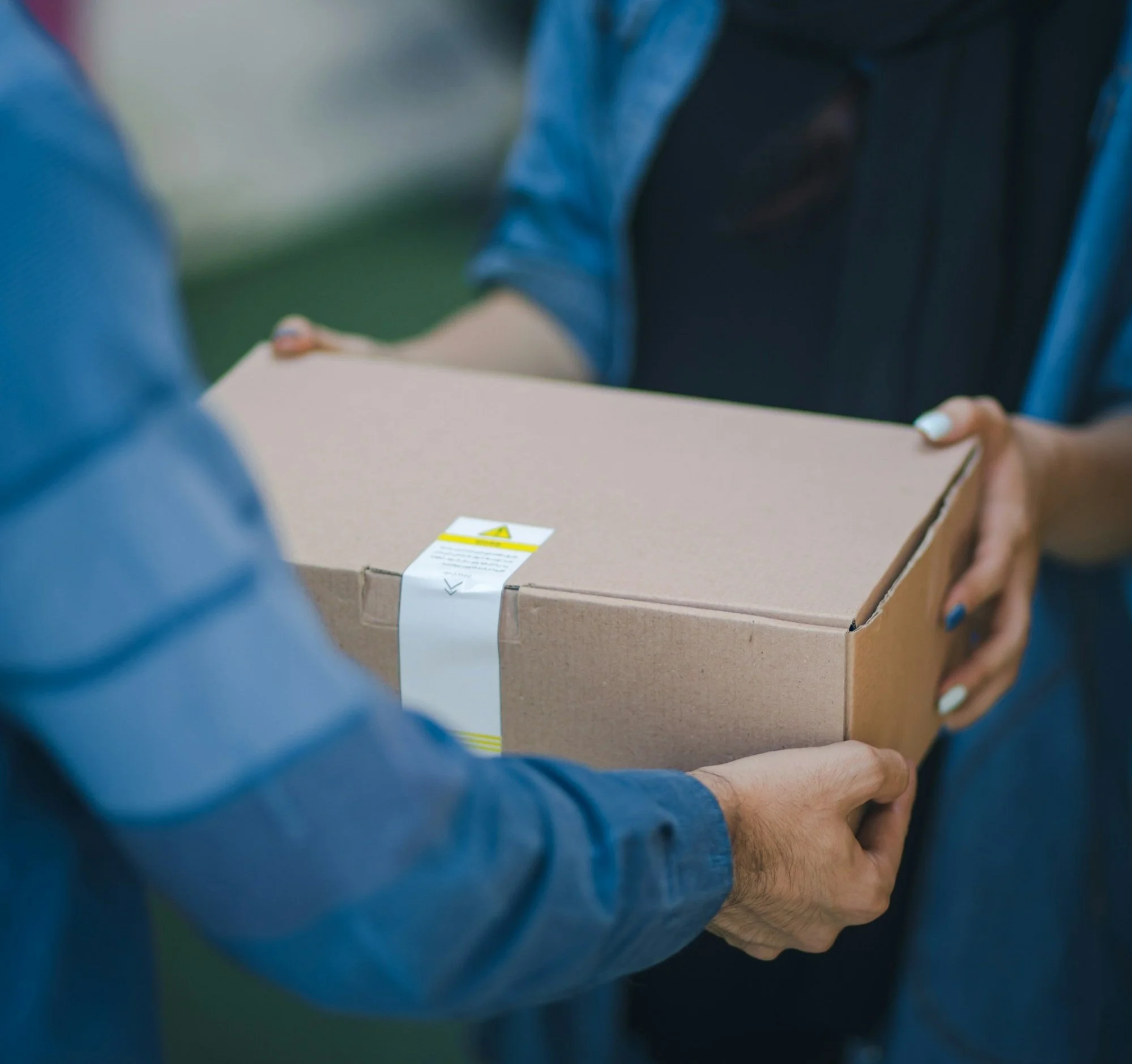 Two people exchanging a cardboard box, one person wearing a blue jacket and the other with painted nails.