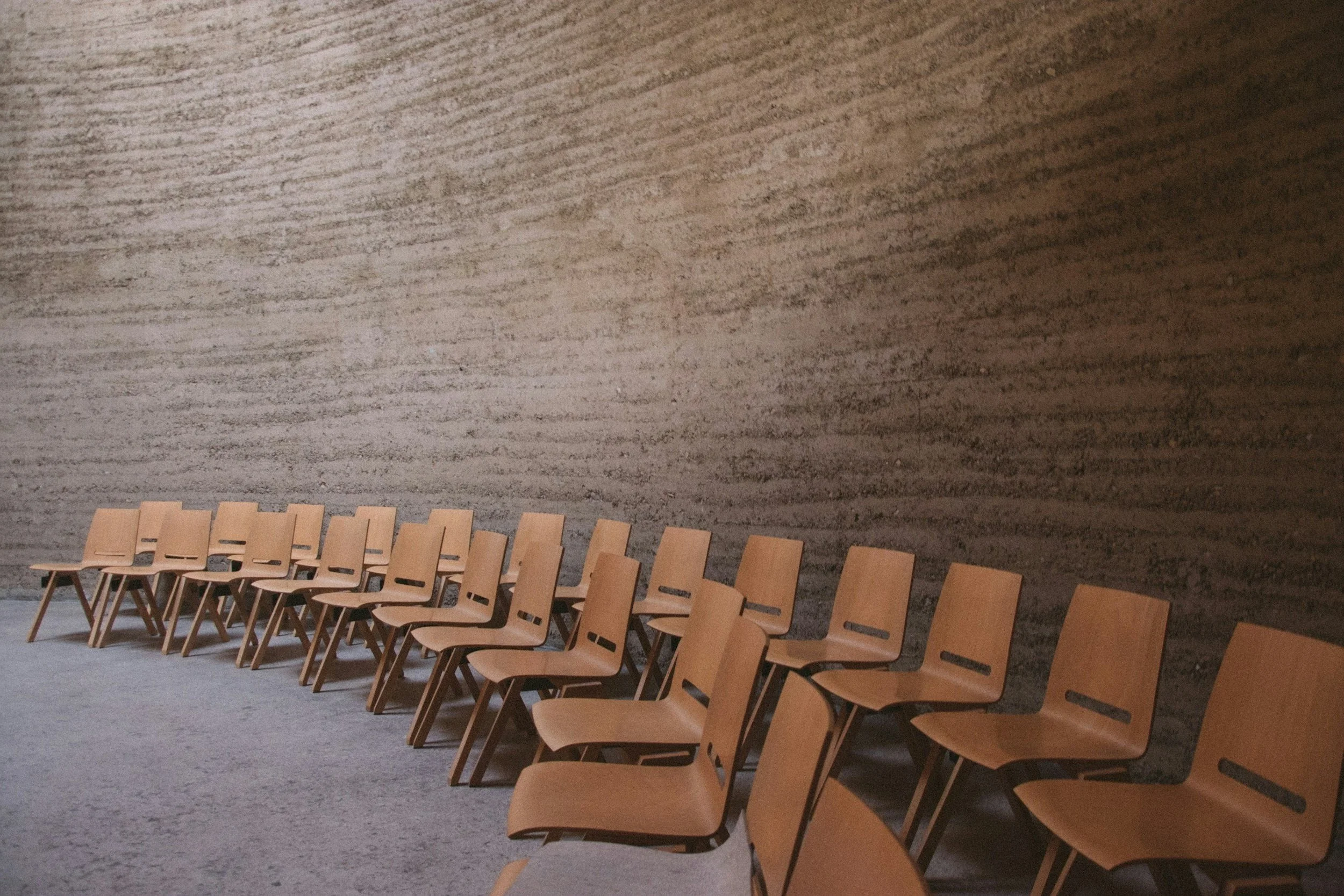 A row of wooden chairs arranged along a textured wall in an empty waiting area or hallway.