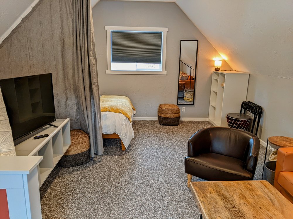 Looking from the lounge area towards the bedroom area with bedroom curtain divider open. In the corner are a wardrobe, luggage racks, poofs, and extra blankets. 