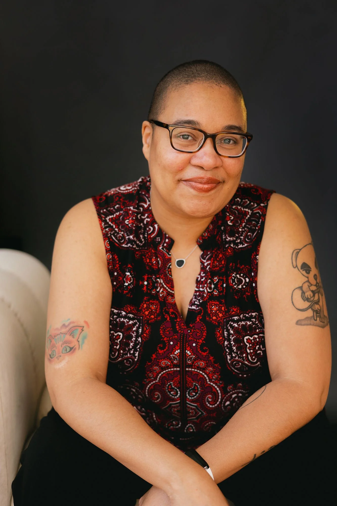 Robyn sits on a cream colored chair, her arms folded in front of her and elbows resting on her legs. She smiles warmly at the camera. She's wearing a sleeveless, red and black paisley shirt so that her arm tattoos are visible.