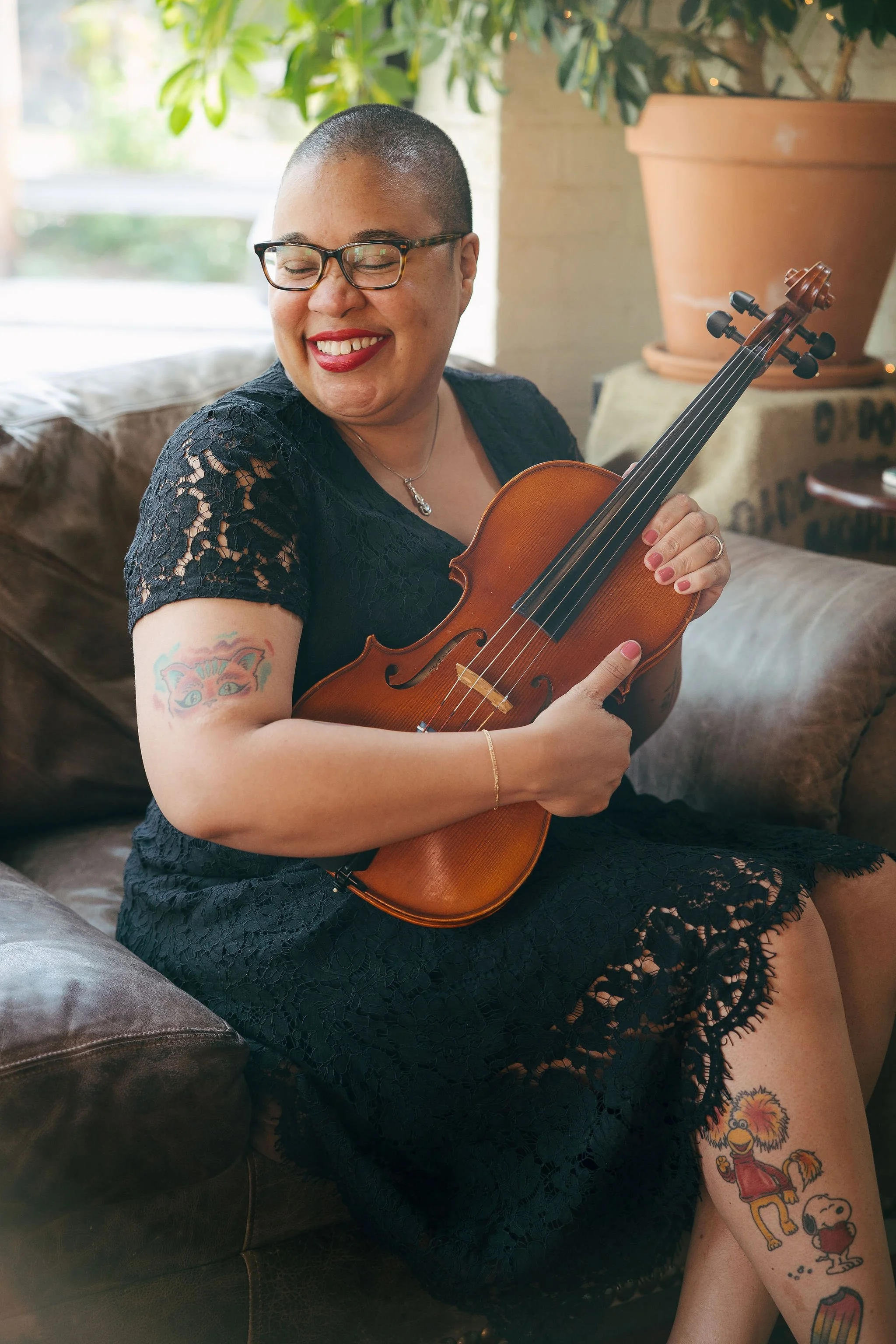 Robyn sits on a couch, smiling with her eyes closed. She holds her viola in front of her, at an angle, and wears a black dress with lacey details. You can see her Cheshire cat tattoo on her right arm and several more tattoos on her right leg.