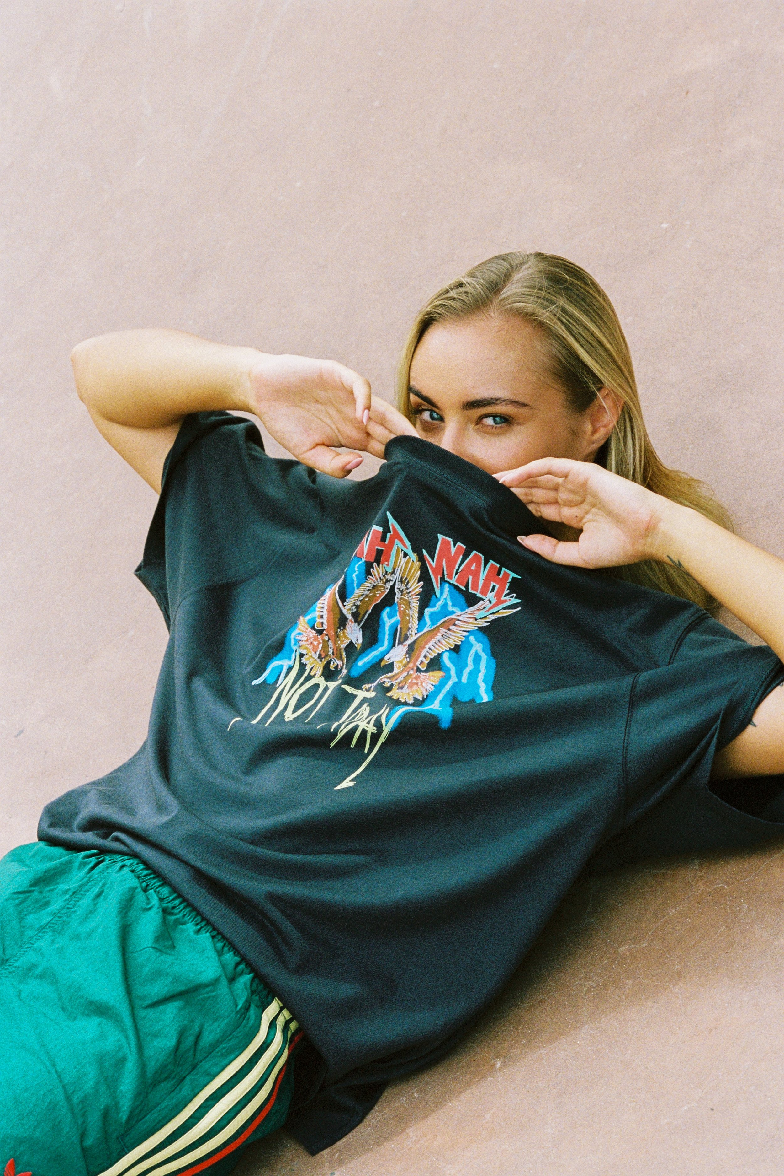 A young woman with blonde hair lying on a tennis court, partially covering her face with a black T-shirt featuring a colorful eagle and lightning design with the text 'THRASHER NOT THA'.