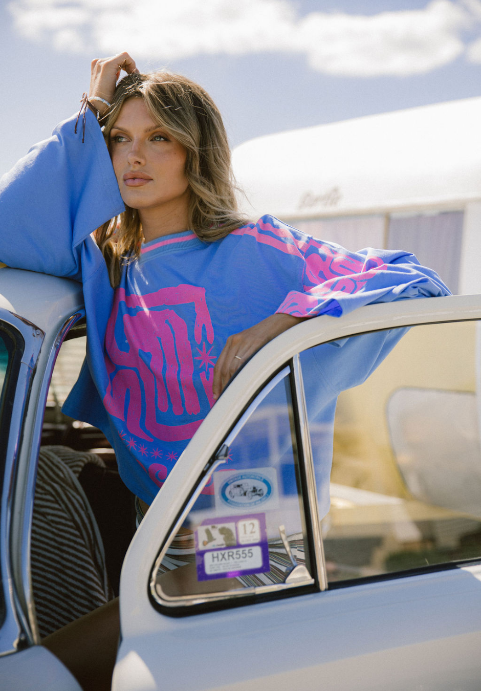 A woman with blonde hair leaning on the open window of a vintage car. She is wearing a blue sweatshirt with pink and purple graphic designs. The background shows a partly cloudy sky and some buildings or trailers.