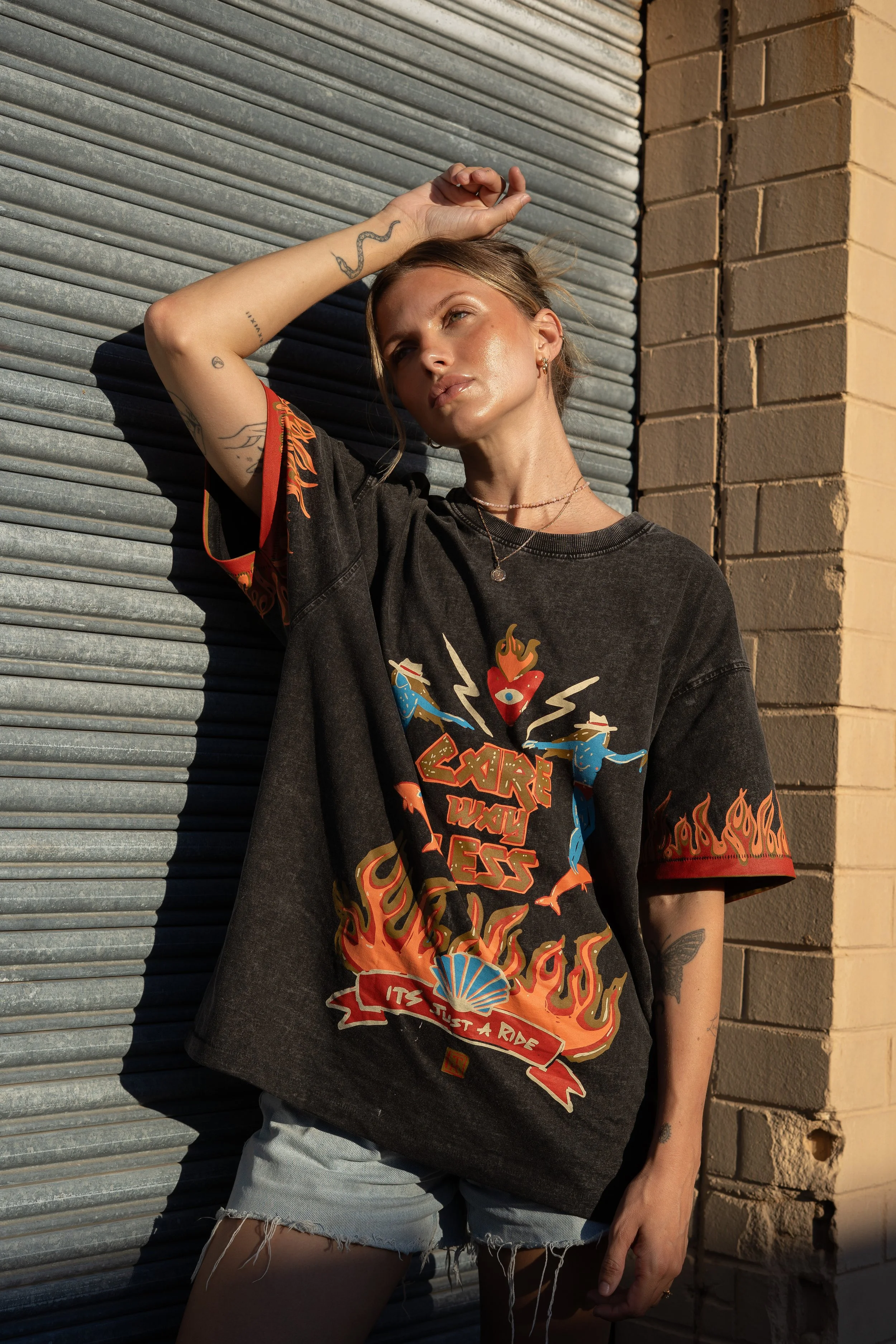 A young woman with tattoos and earrings, wearing a graphic black t-shirt and ripped denim shorts, is leaning against a metal garage door and brick wall in sunlight.
