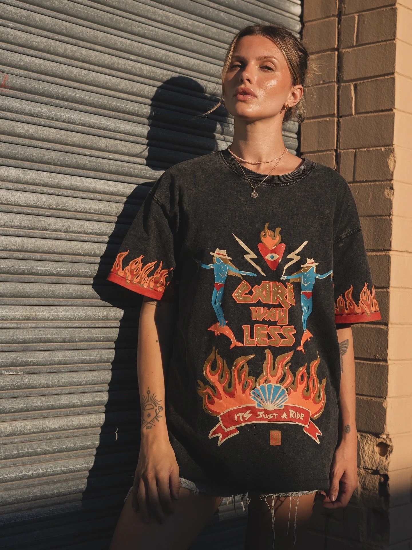 A young woman with light skin and brown hair stands outdoors against a metal garage door and brick wall. She is wearing a black graphic t-shirt with colorful flames, dancing women, a heart, and text that reads "CARRY ME LESS, IT'S JUST A RIDE." The sunlight is casting shadows, and she has tattoos on her left arm.