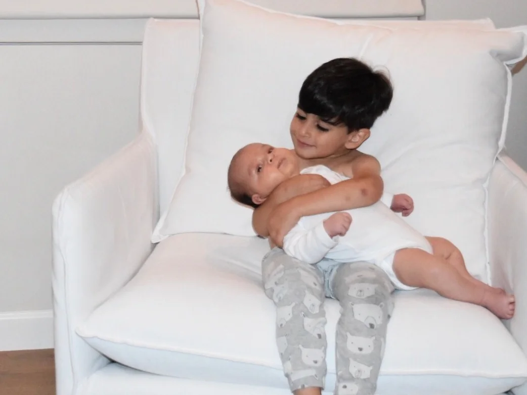 El’s middle son Leo sitting on a white chair holding newborn baby brother Max
