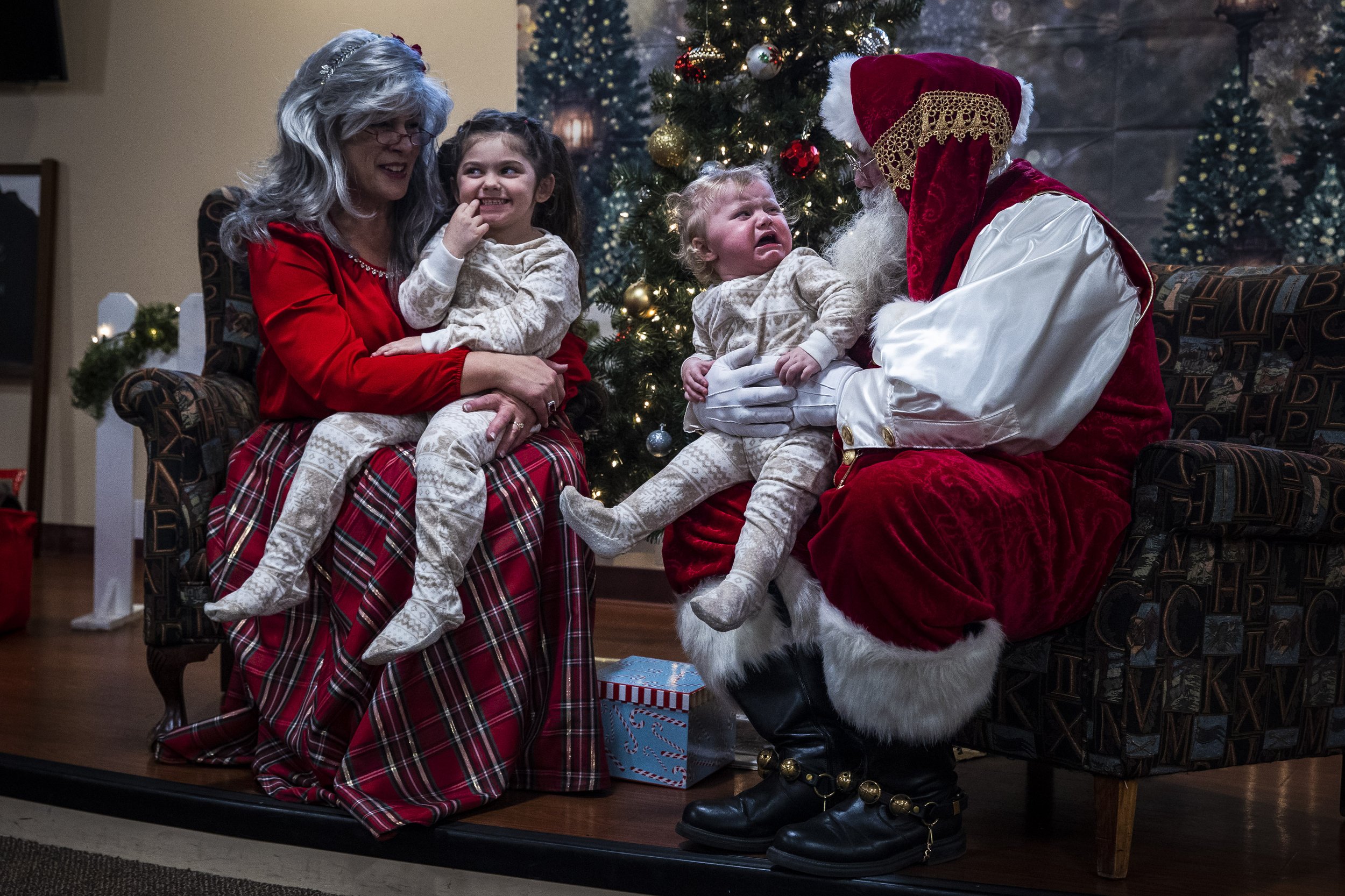  One-and-half-year old Adalynn Knapp cries after realizing that her mother placed her onto the lap of Santa Claus, portrayed by Rick Banks, on December 10, 2022. Her five-year-old sister Alayla Panzino sits happily with Mrs. Claus, Anne Banks.  Suiti