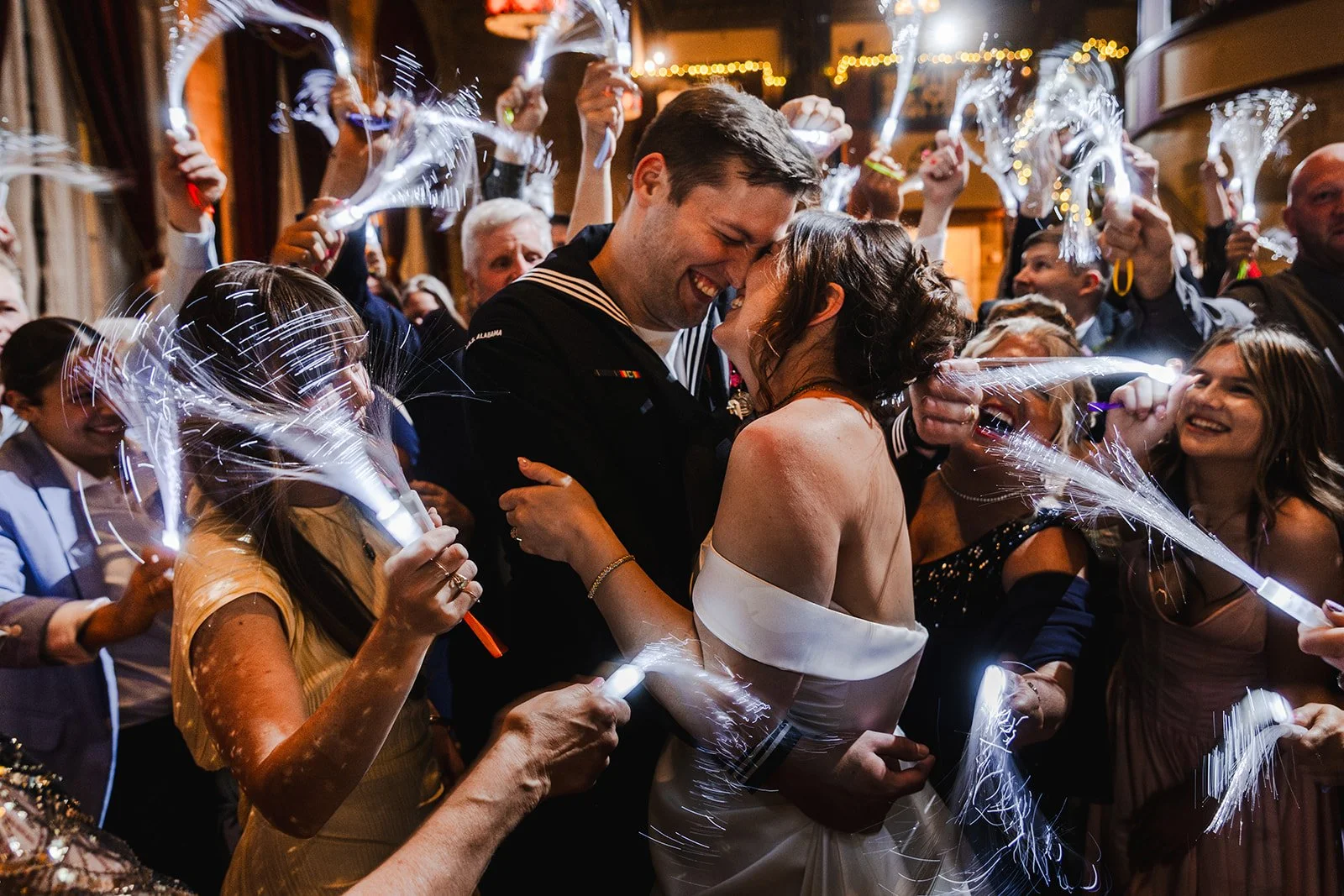 A man and woman in wedding attire are embracing and smiling at each other amid a crowd celebrating at a wedding reception. Guests are holding sparklers, creating bright streaks of light in the festive indoor setting.