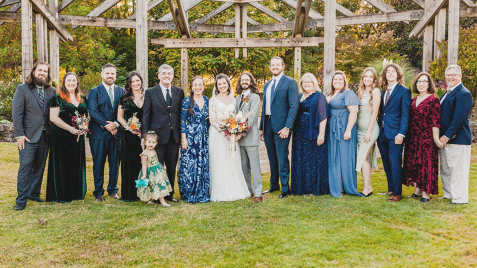 A group of people at a wedding, including the couple in the center, standing outdoors on grass with wooden structure and trees in the background.